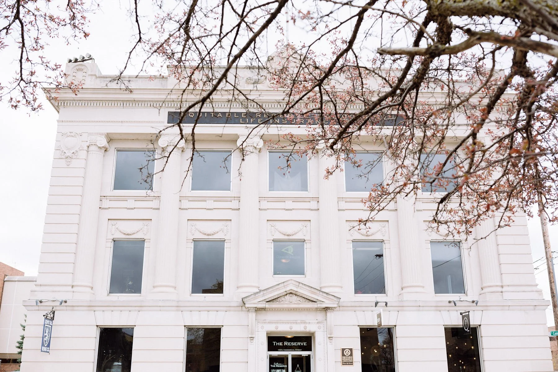 White building with classical architecture, including columns and decorative elements, with a sign that reads 'The Reserve' above the entrance, and a tree with pink blossoms partially visible in the foreground.