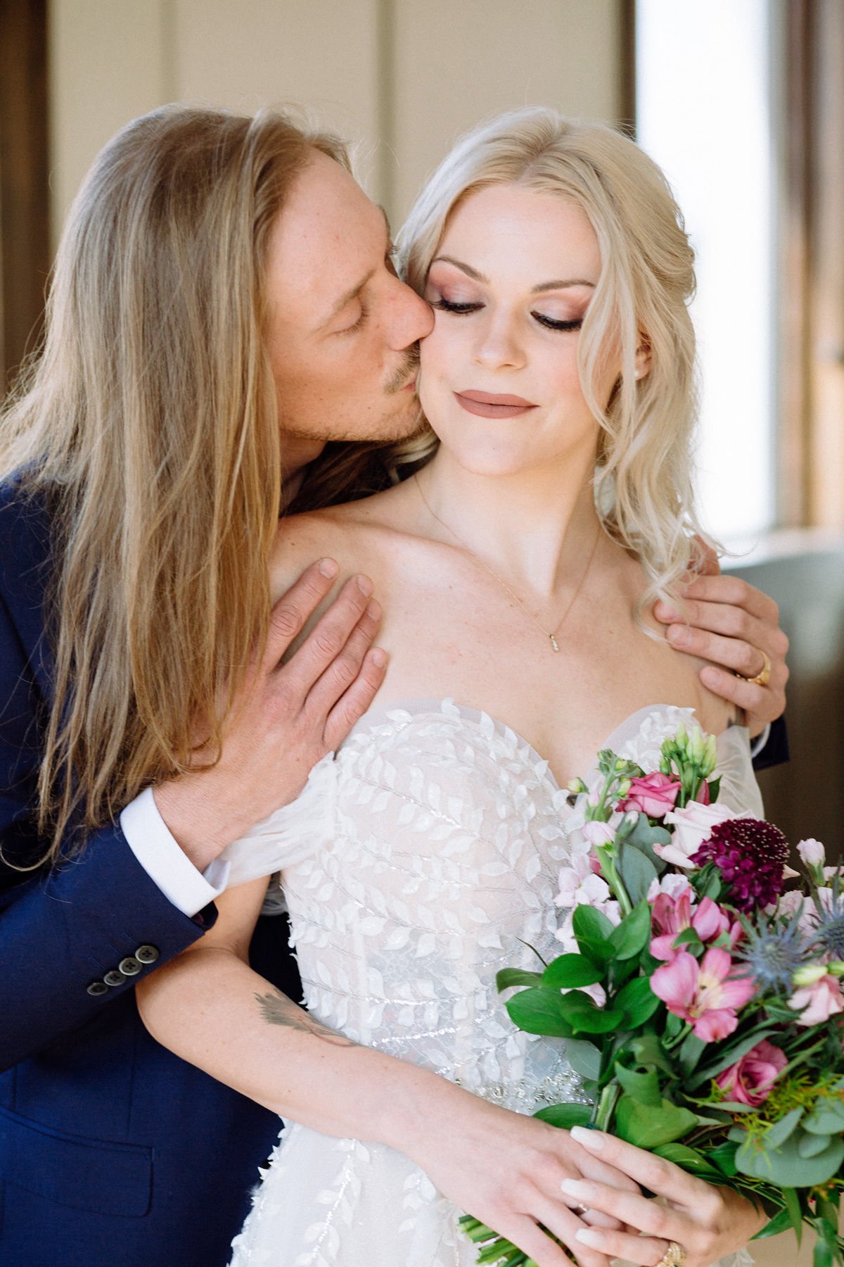 A groom kisses a bride on the cheek while holding her shoulders. The bride holds a bouquet of pink and purple flowers and wears a white wedding dress. They are indoors with a neutral background.