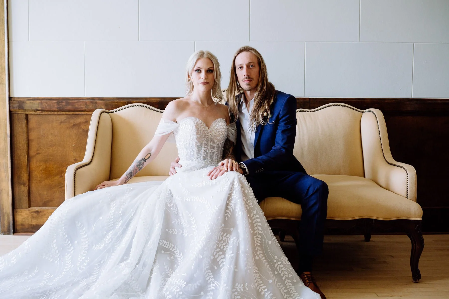 A bride and groom sitting together on a beige vintage sofa, indoors. The bride is wearing a white off-shoulder wedding gown with lace details, and the groom is in a dark blue suit with a white shirt. They are holding hands, with neutral expressions.