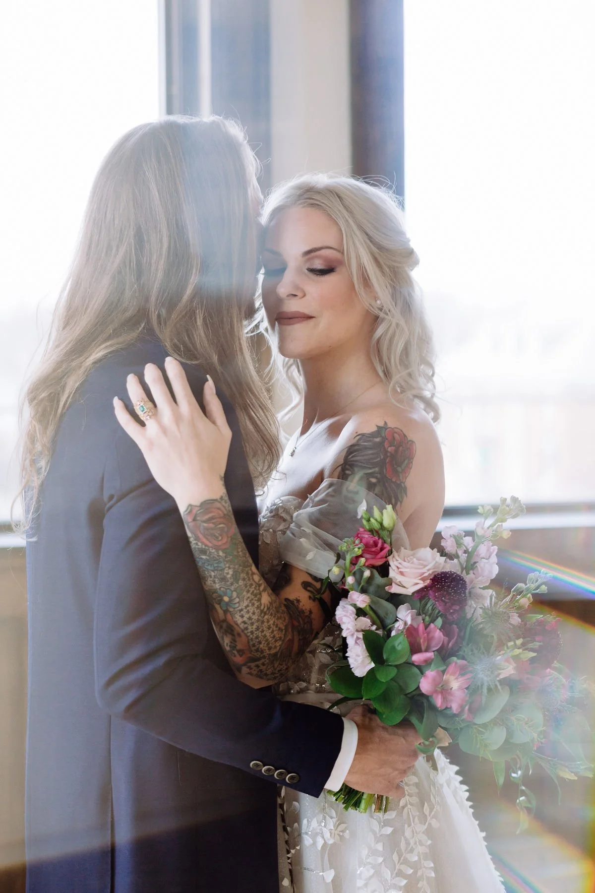 A couple dressed in wedding attire, embracing and holding a bouquet of flowers near a bright window.