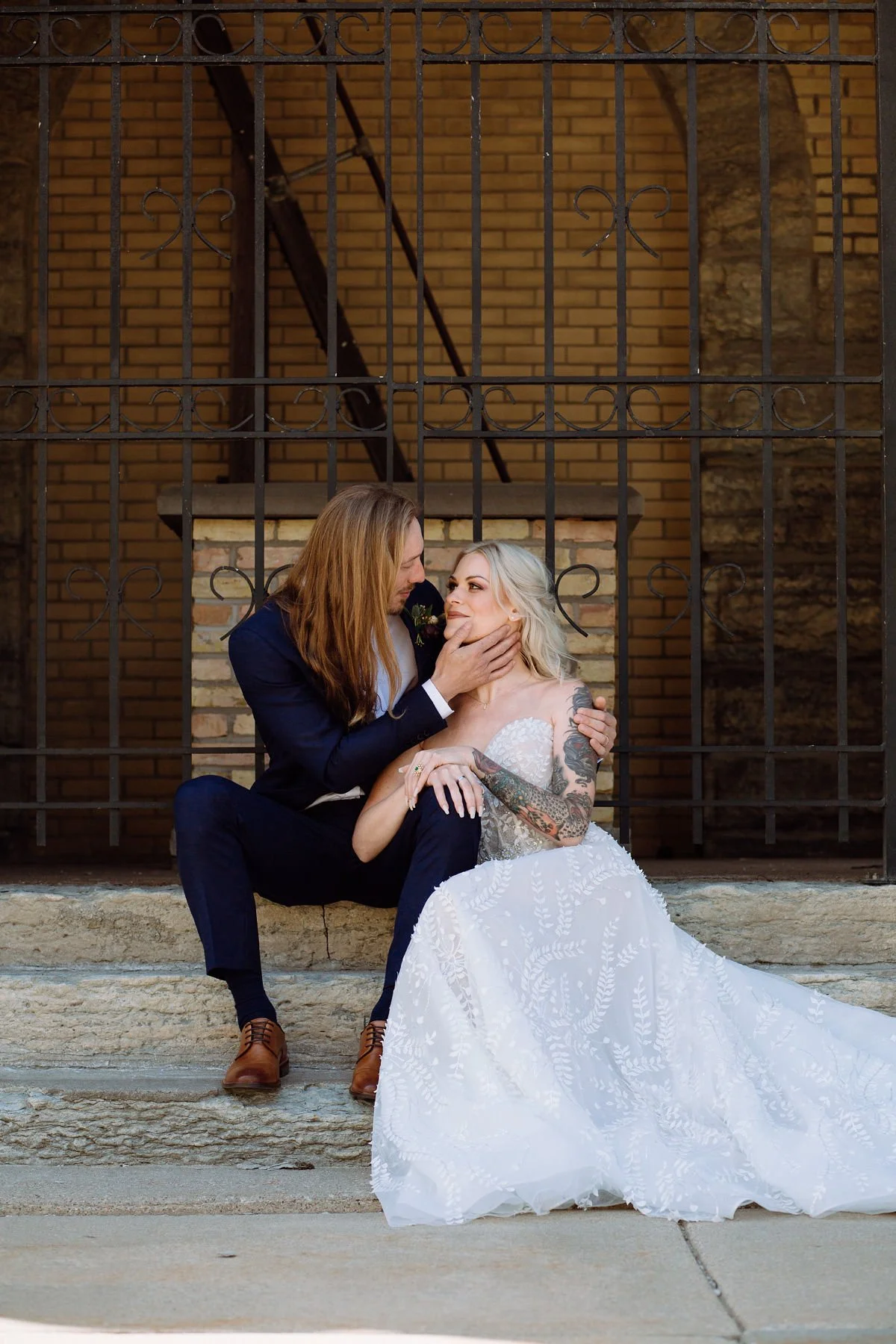 A couple dressed in wedding attire sitting on stone steps behind a wrought iron fence, with a brick wall in the background. The man has long hair and is gently holding the woman's face while she looks at him.