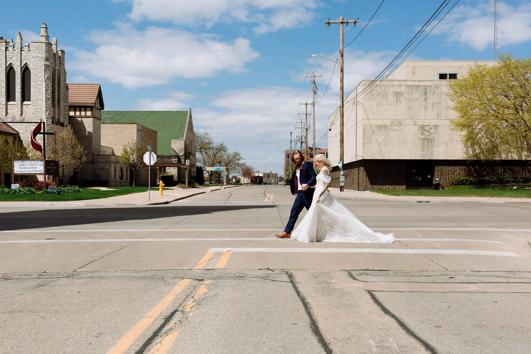A bride and groom crossing a street in an urban area on their wedding day, with buildings and utility poles in the background.