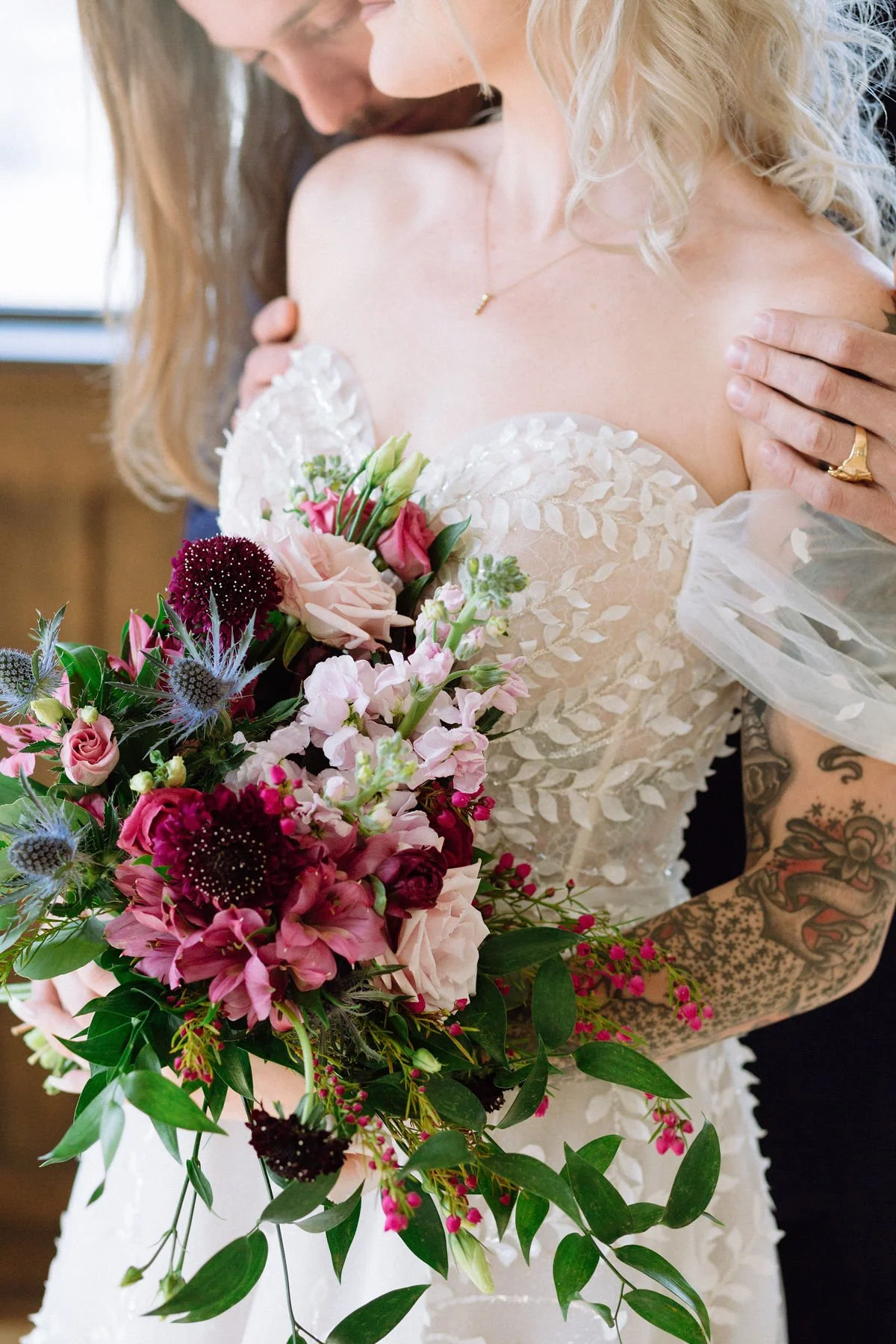Close-up of a bride holding a bouquet of pink, purple, and white flowers, with a person embracing her from behind, both dressed in wedding attire.