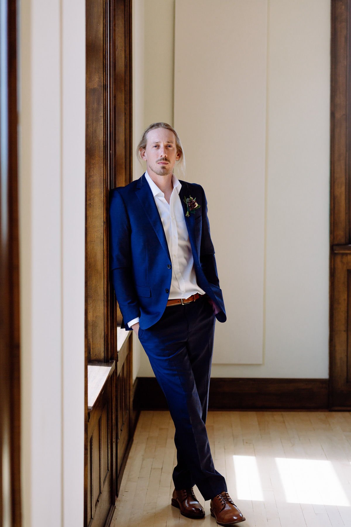 A man with blond hair and a light beard leaning against a wooden wall. He is wearing a dark blue suit, white dress shirt, brown belt, and brown shoes. He has a boutonniere attached to his left lapel and is in a room with cream-colored walls and natur