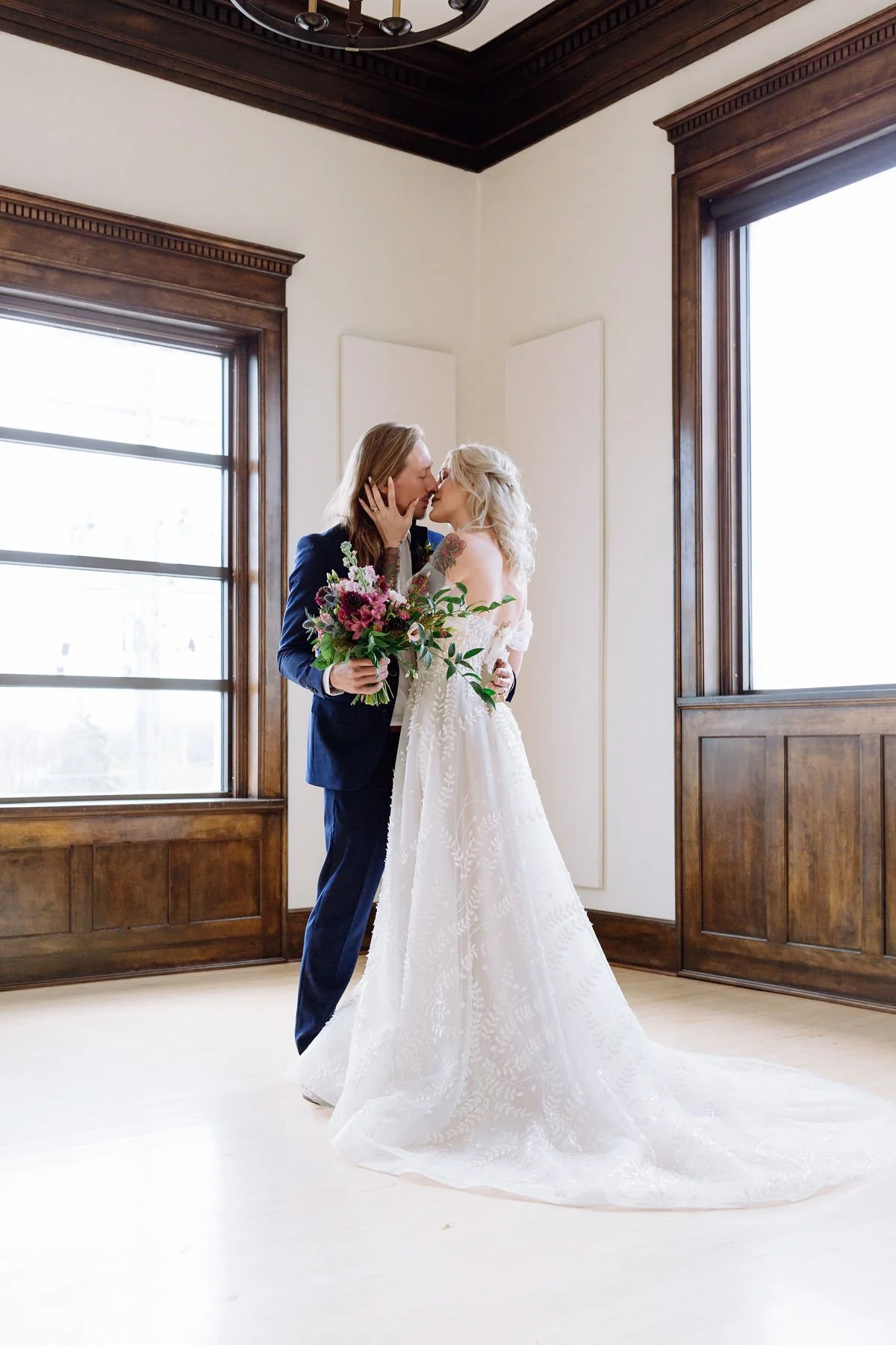 A couple, dressed in wedding attire, kissing inside a room with large windows and wooden trim. The bride holds a bouquet of flowers and wears a white wedding gown. The groom is dressed in a dark suit.