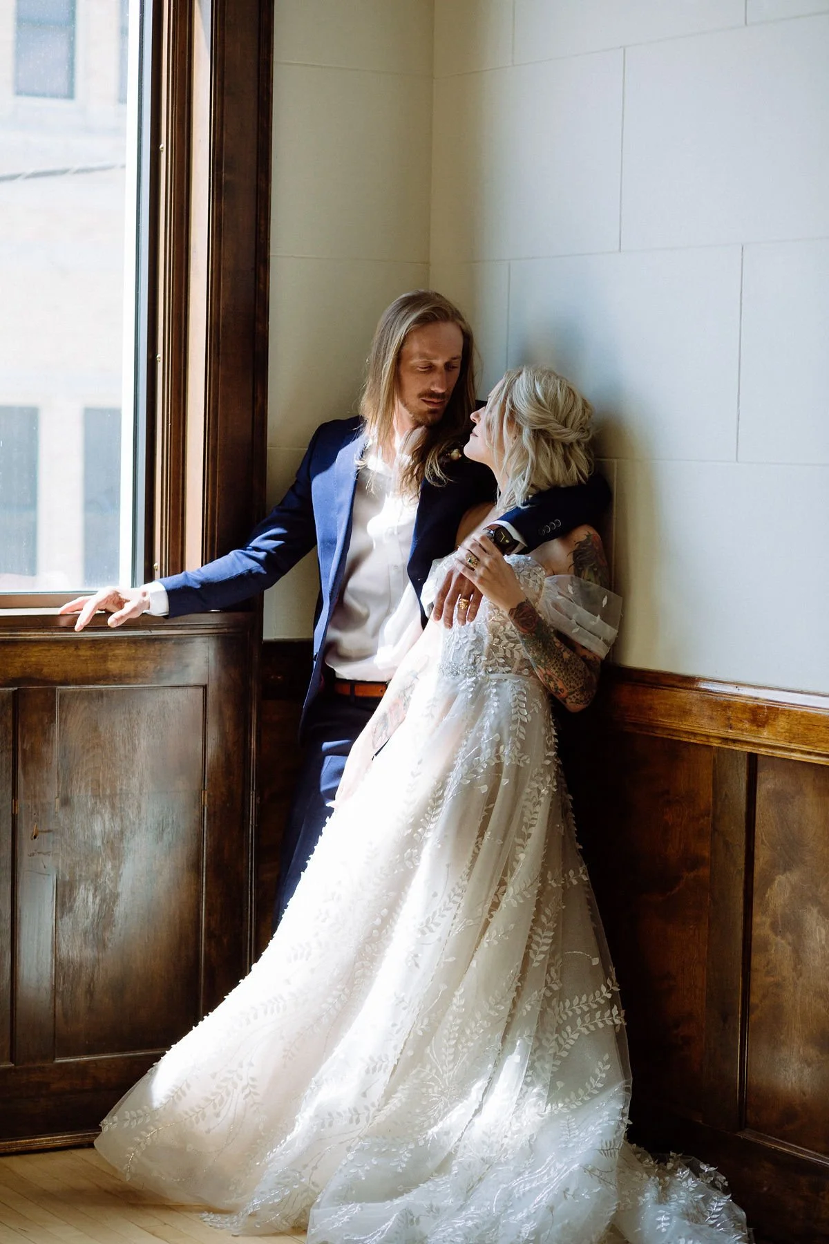 A couple dressed in wedding attire, the woman in a white lace gown and the man in a dark suit, sharing an intimate moment by a window with sunlight streaming in.