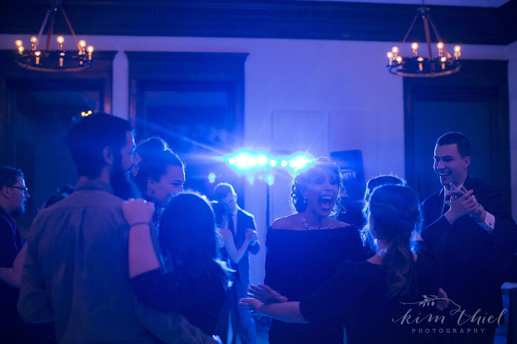 People dancing and celebrating at a party with blue lighting and chandeliers in the background.