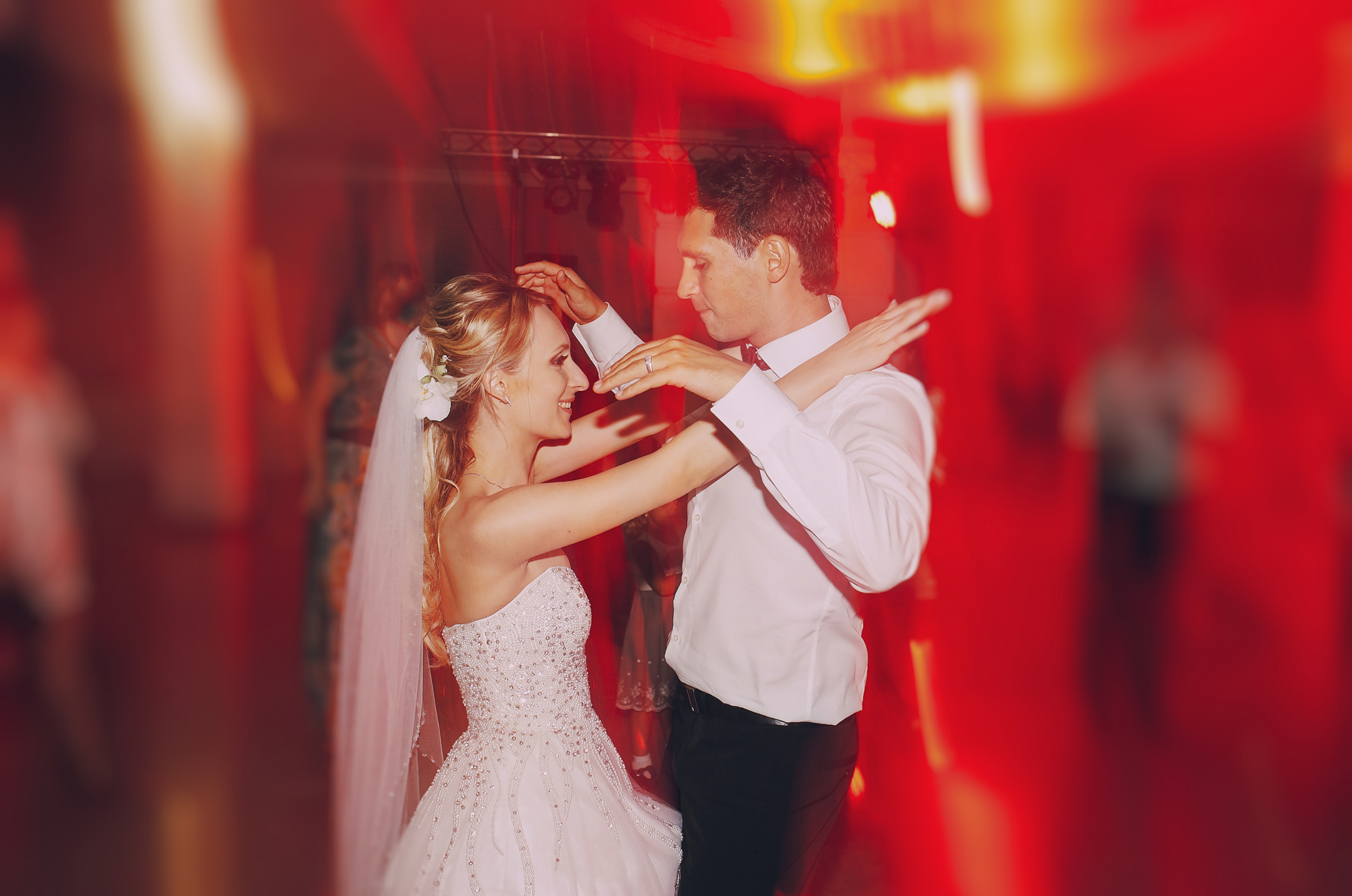 A newlywed couple sharing a dance at their wedding reception, with red lighting and blurred guests in the background.