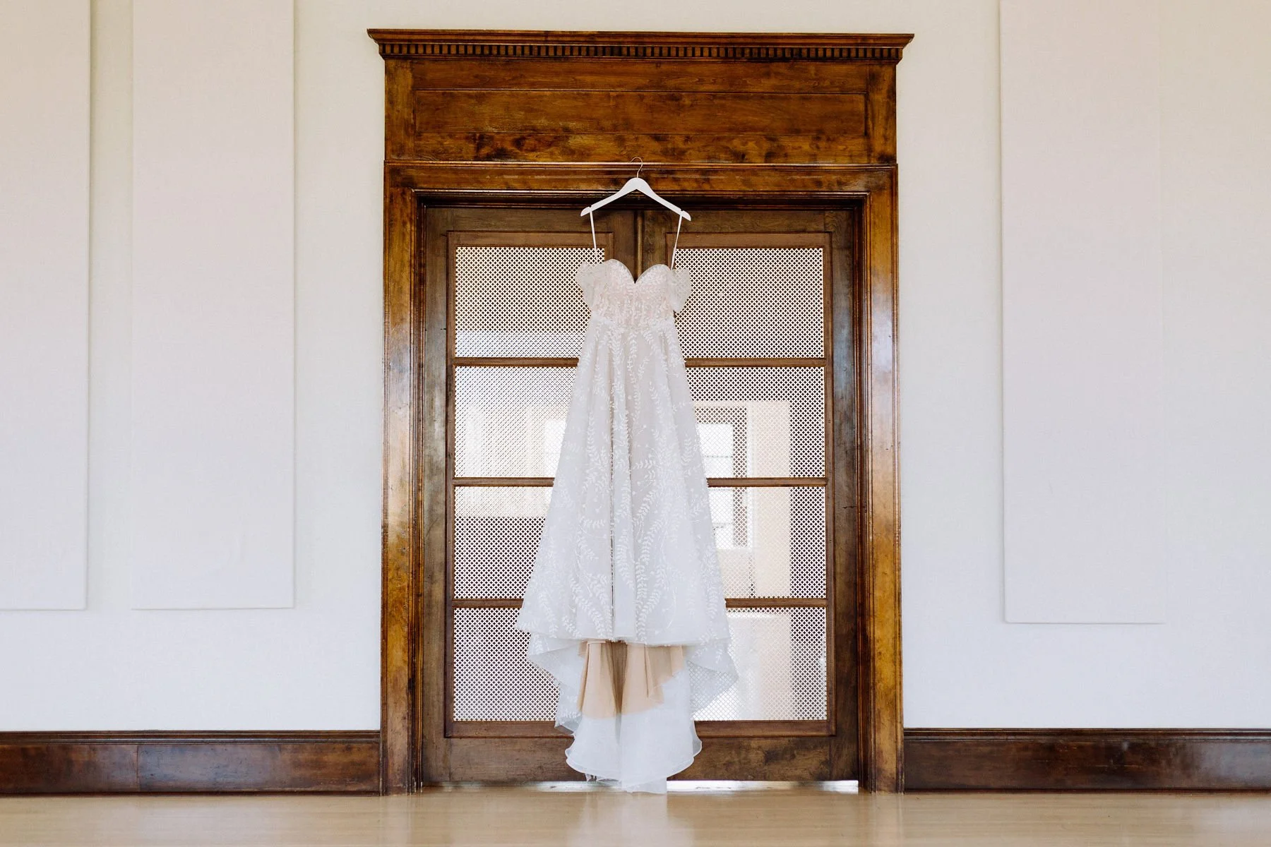 White wedding dress hanging on a wooden door with glass panes and a background of a bright room.