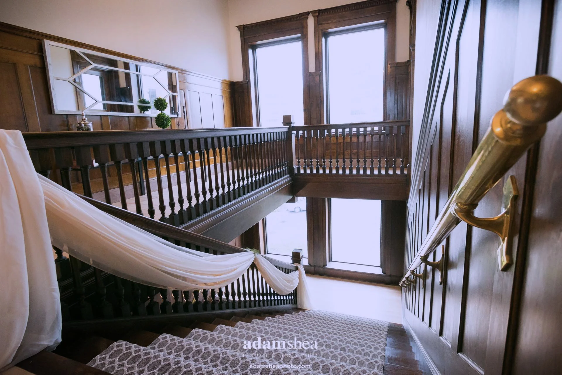 Interior view of a vintage wooden staircase with a banister, large windows letting in natural light, wood-paneled walls, framed mirror, decorative plants, and a draped fabric over the banister.