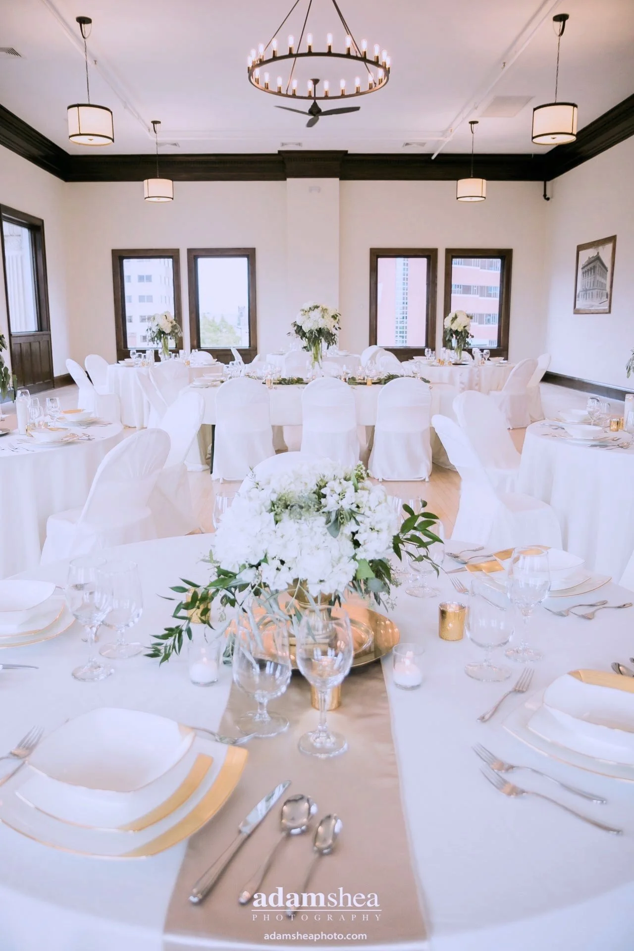 Elegant banquet hall decorated for a wedding or special event with round tables covered in white tablecloths, floral centerpieces, and white chair covers. Large windows provide natural light, and a chandelier hangs from the ceiling.