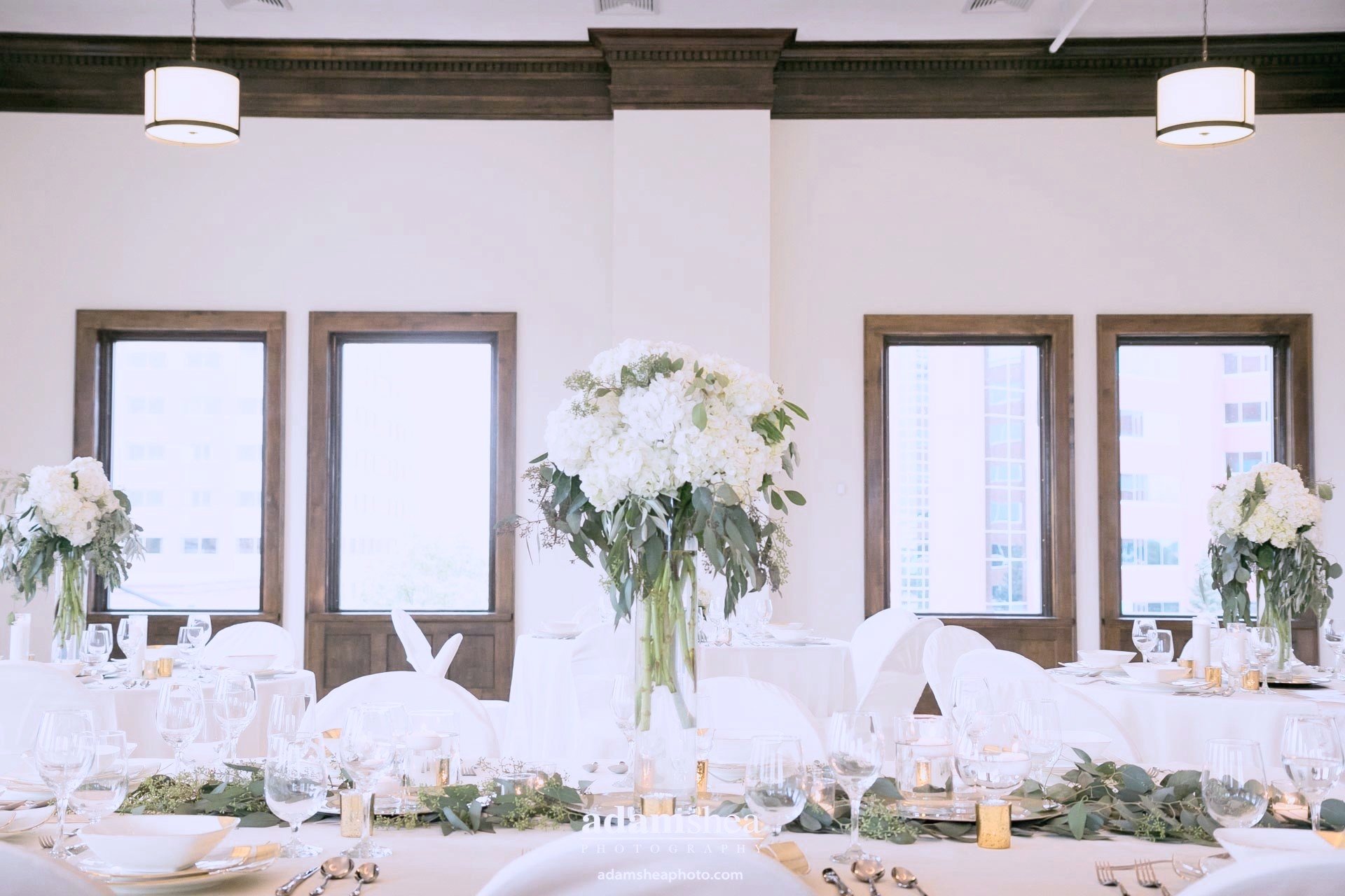 Elegant banquet table set with white tablecloths, floral centerpieces with white flowers and greenery, and neatly arranged glassware, utensils, and plates in a bright room with large windows and modern lighting fixtures.