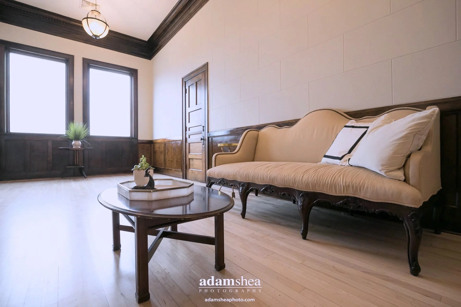 A bright living room with two large windows, a beige vintage sofa, a wooden coffee table with a decorative tray, and a small plant on a stand near the window.