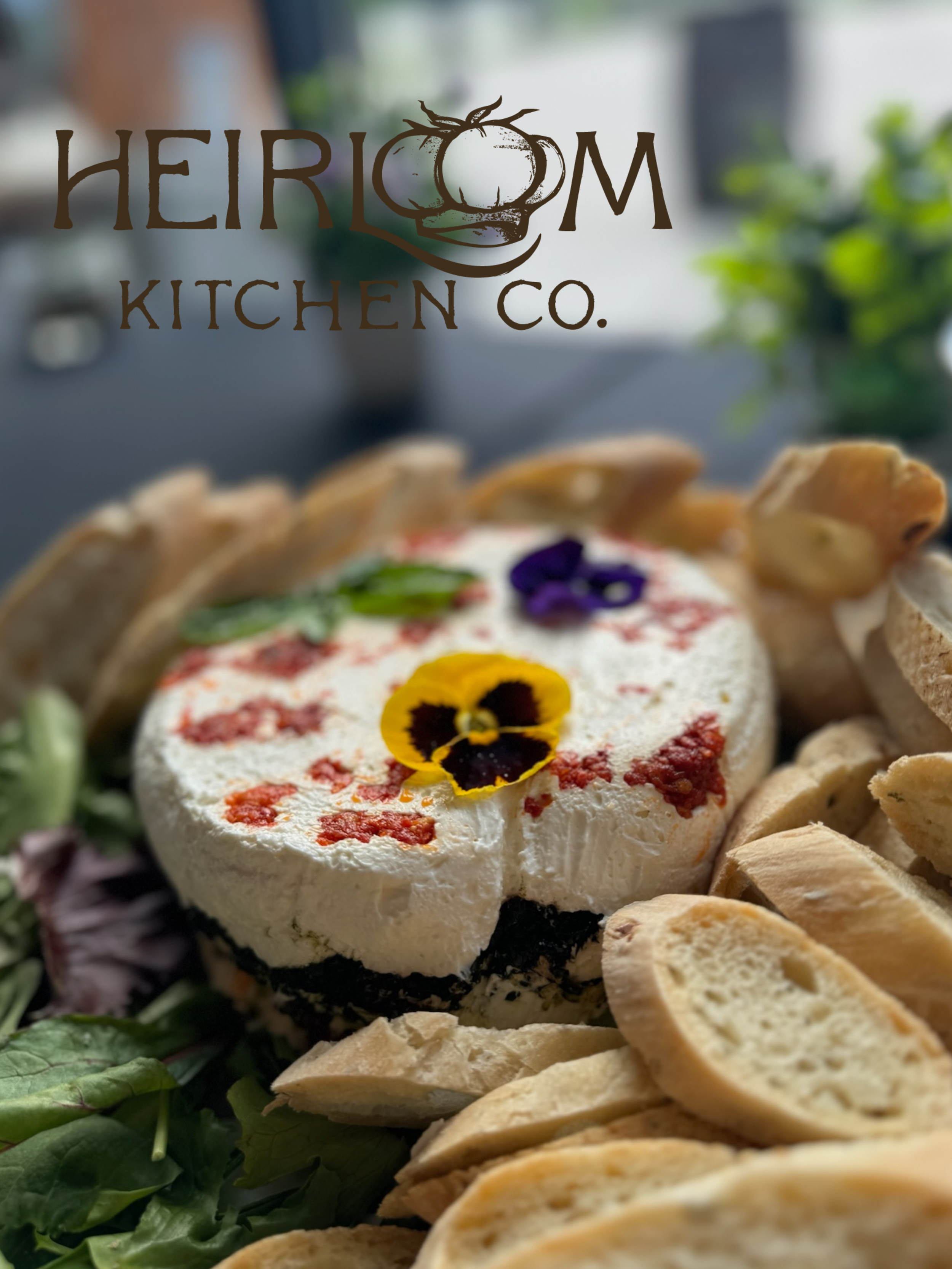 Cheesecake topped with edible flowers, surrounded by biscotti and green leaves, with a logo for Heirloom Kitchen Co. in the upper part of the image.