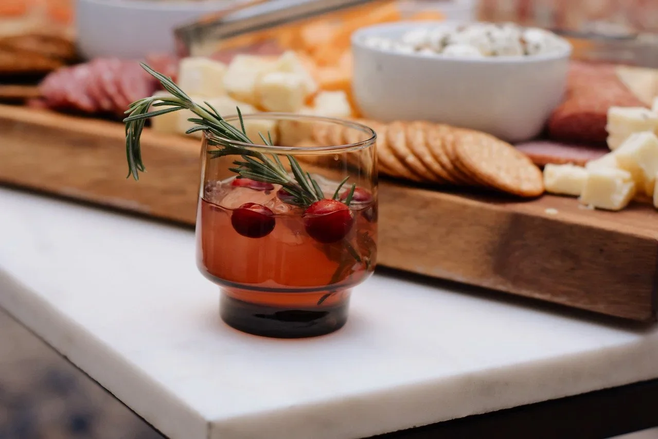 Glass of red beverage with cranberries and rosemary, served on a white surface, with a cheeseboard of cheese, crackers, and cured meats in the background.