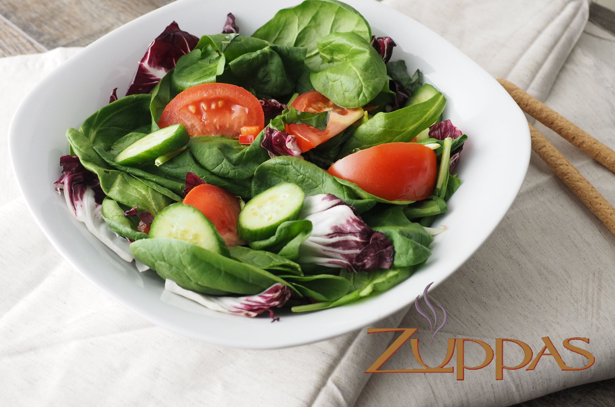 Fresh garden salad with lettuce, spinach, tomato, cucumber, and radicchio in a white bowl on a beige cloth with breadsticks nearby.