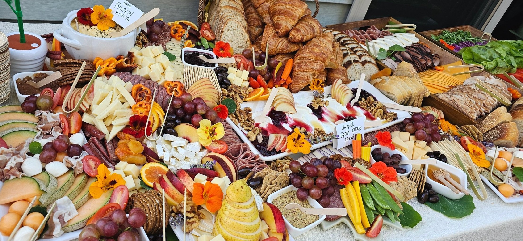 A large spread of foods including assorted cheeses, sliced meats, fresh fruits, bread, croissants, crackers, spreads, and garnishes arranged on a table, with labels indicating items like whipped goat cheese spread and savory pumpkin ricotta.