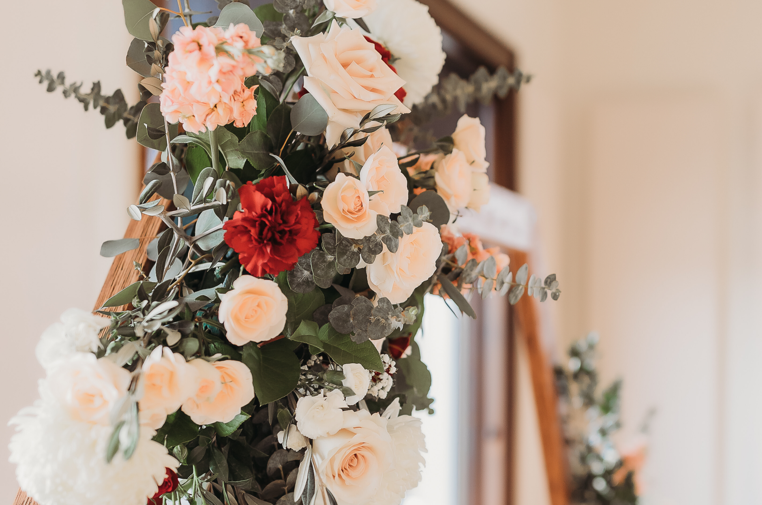 Close-up of a flower arrangement with cream, pink, and red roses, white carnations, peach-colored flowers, eucalyptus leaves, and greenery, attached to a wooden frame or decorative stand.