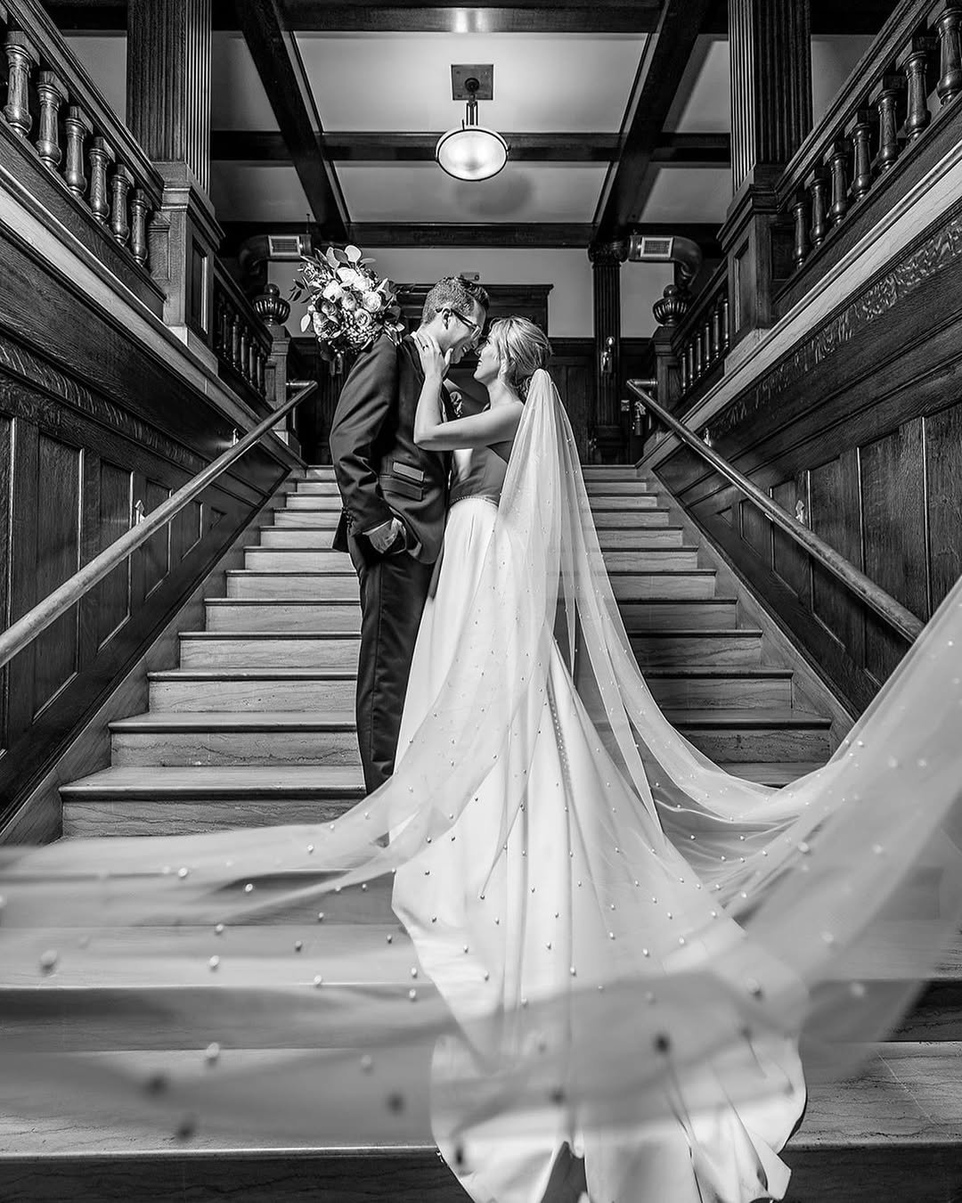 Black and white photo of a bride and groom on a staircase. The bride is in a white gown with a long train and veil, holding the groom's face. The groom is in a suit, and they are gazing into each other's eyes.