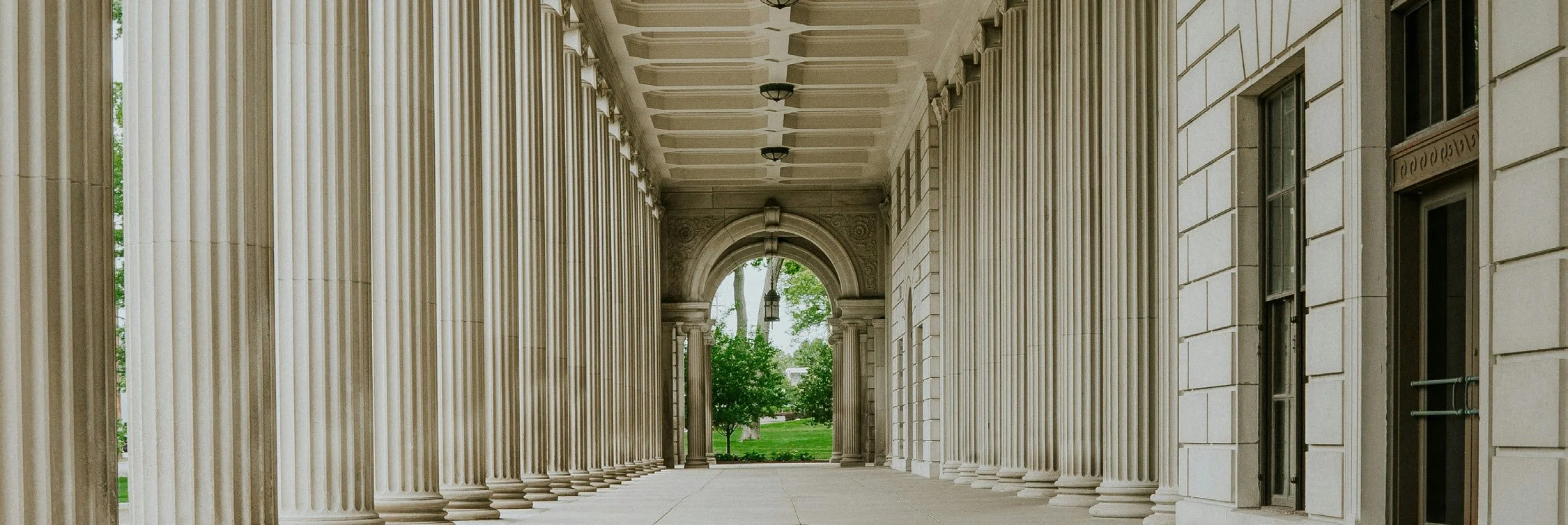View of a classical building with tall, white columns along a walkway, leading to an outside area with green trees.