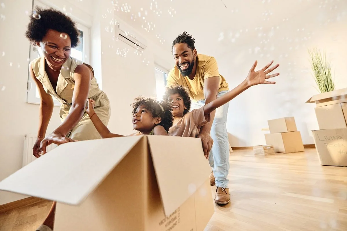A family of four moving into a new home; the parents and two children are smiling and playing with a moving box inside a bright, modern room.
