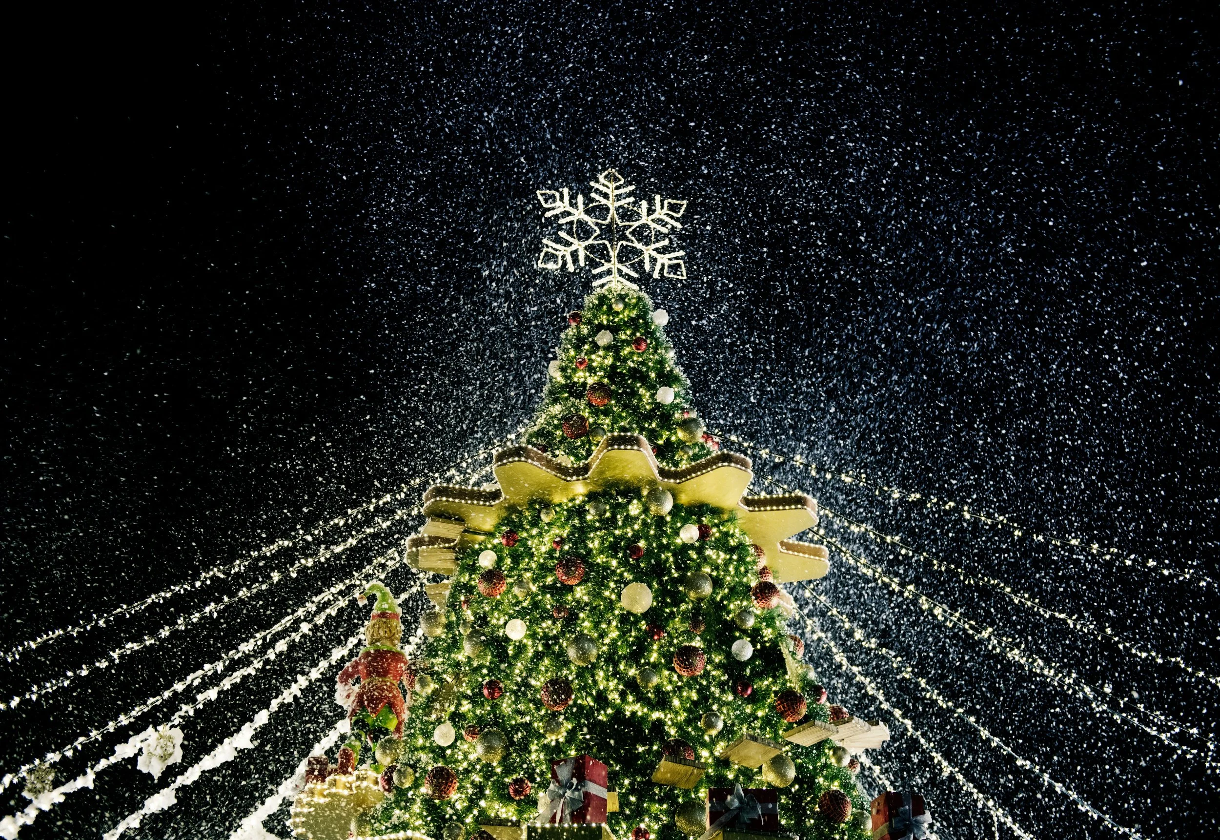 Christmas decorated tree with ornaments and lights, topped with a snowflake-shaped decoration, under falling snow at night.
