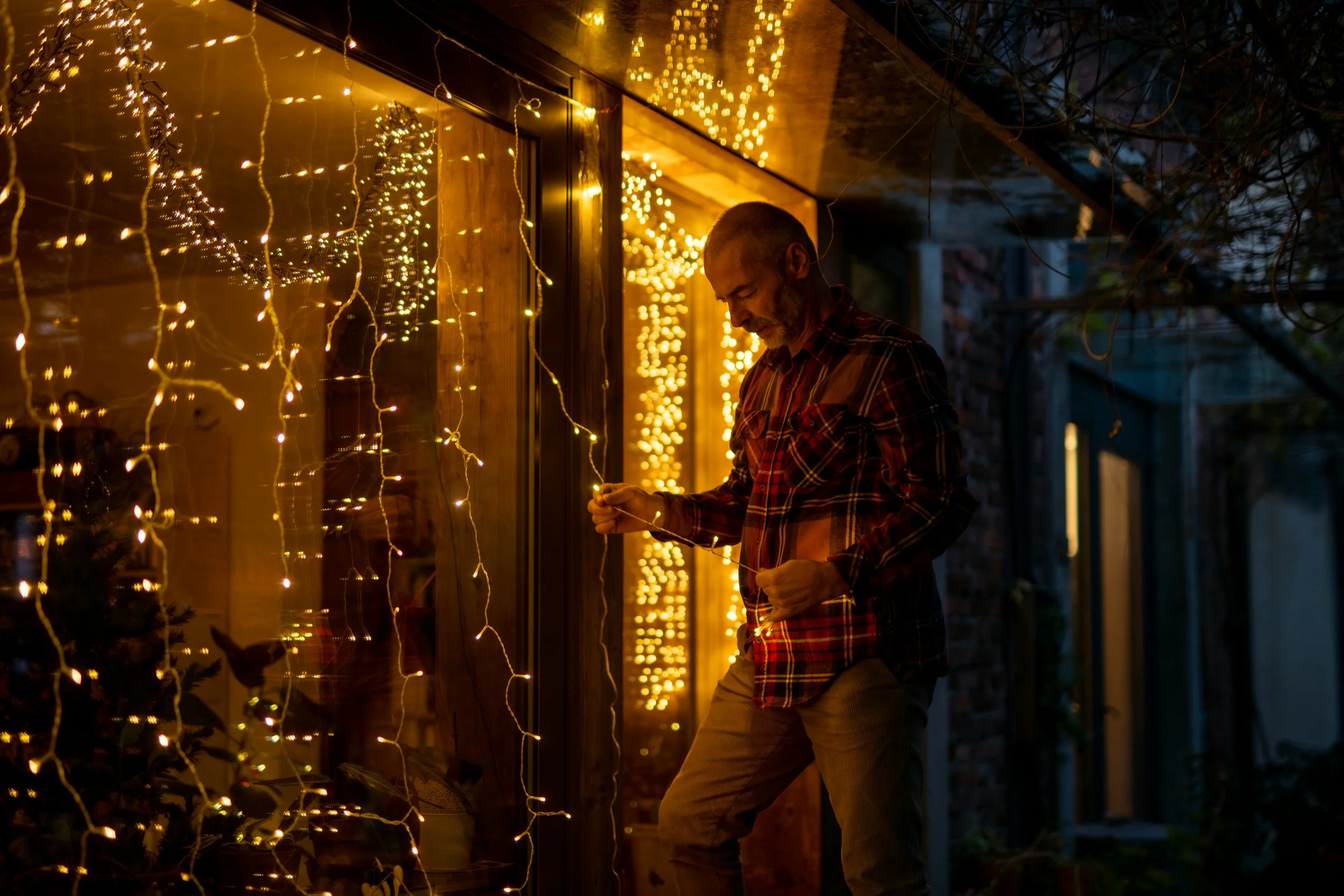 A man in a plaid shirt hanging Christmas lights on a house porch during evening, illuminated with warm yellow string lights.