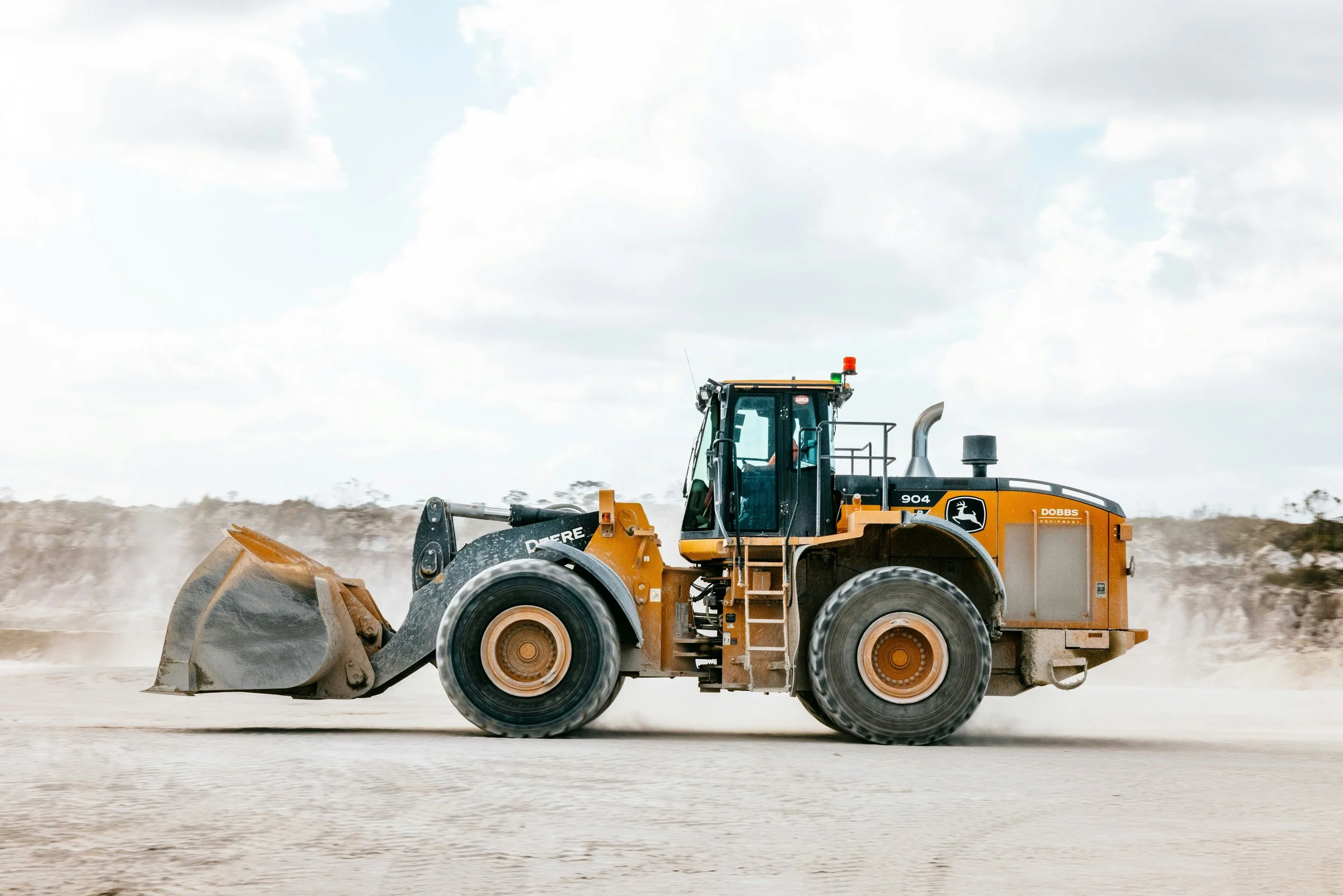 A large yellow construction vehicle, specifically a wheel loader, operating on a dusty construction site with clouds in the sky in the background.