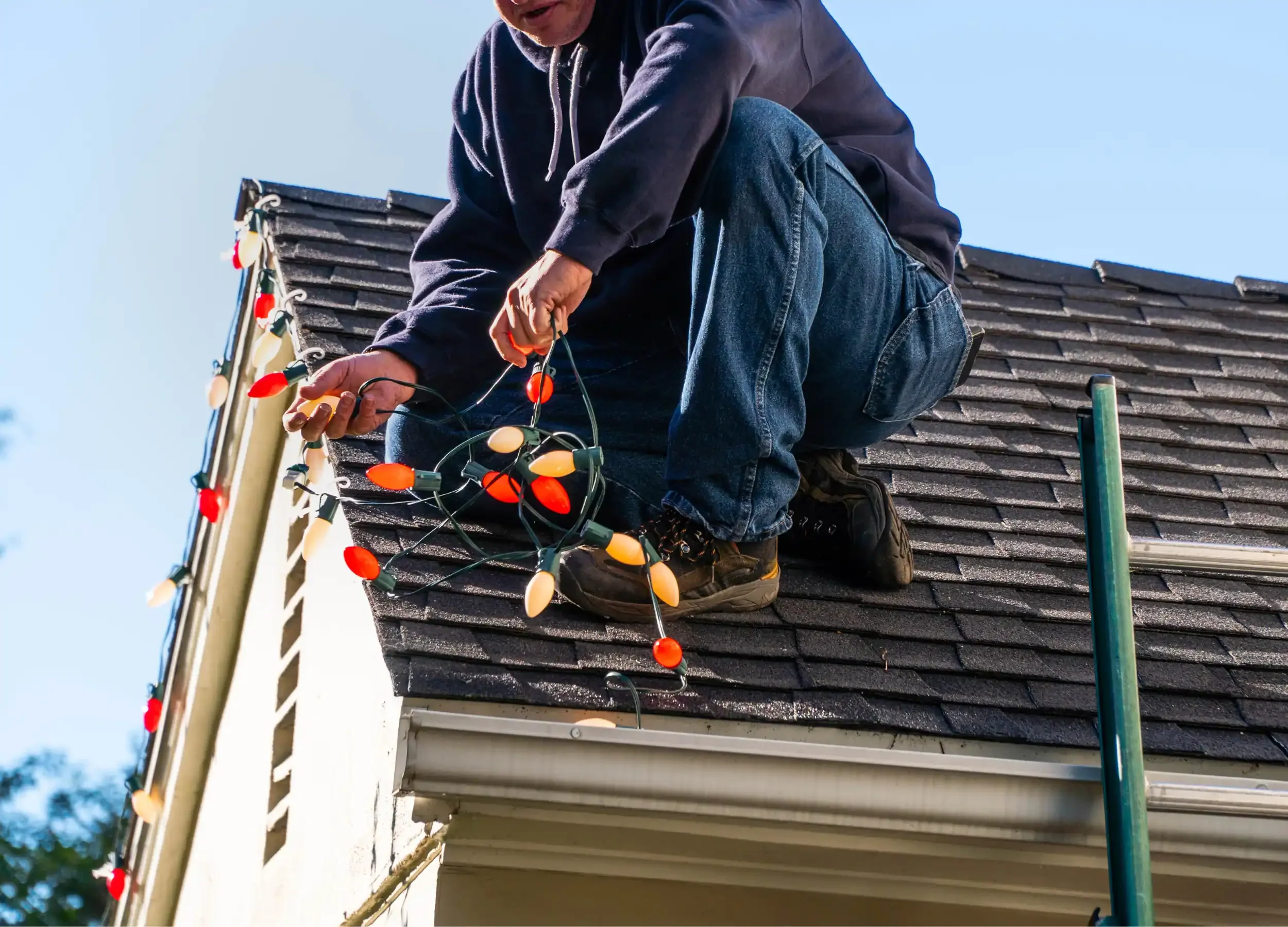 A person on a roof decorating for Christmas with colorful string lights.