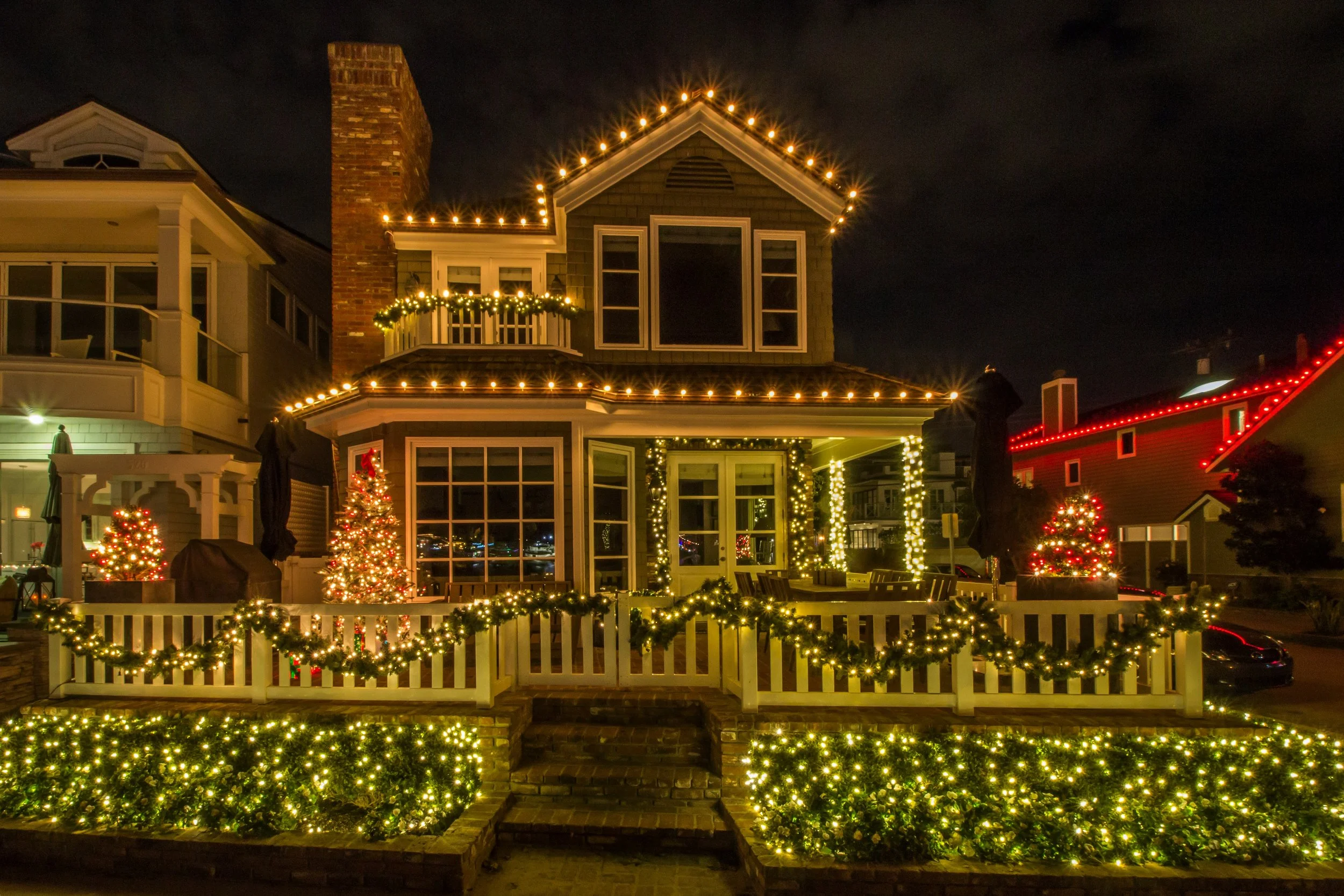 A two-story house decorated with Christmas lights at night, featuring string lights outlining the roof, garland with lights on the porch columns, and Christmas trees with ornaments illuminated outside. The house has a white picket fence decorated with lights.