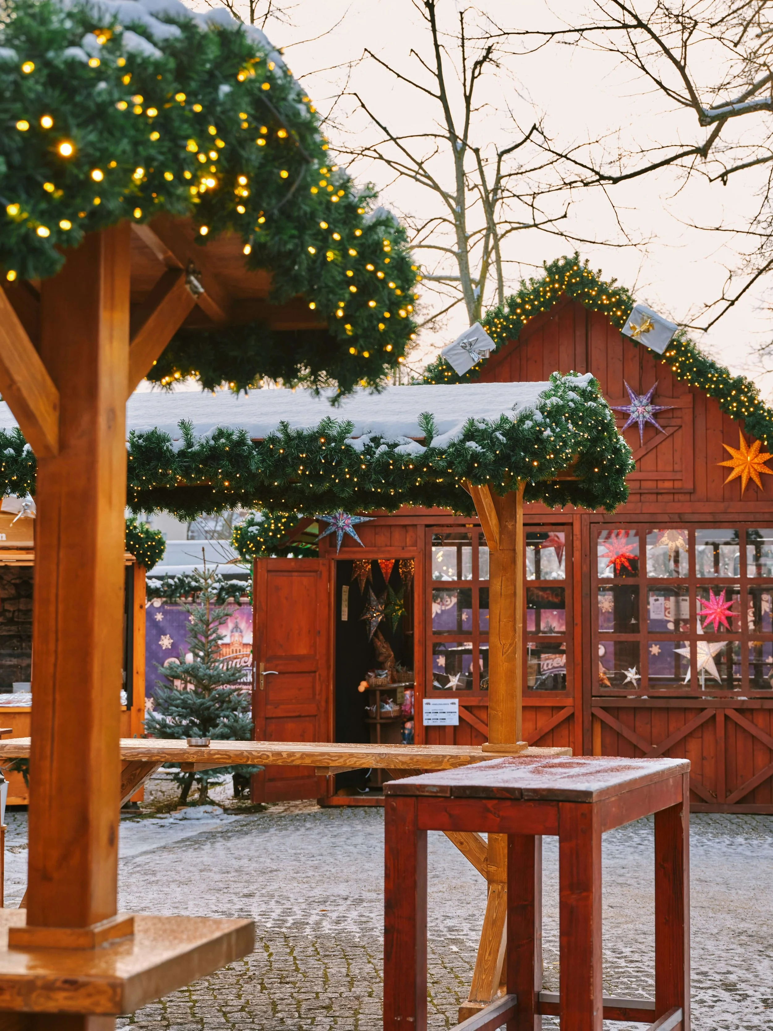 A festive outdoor Christmas market scene with decorated wooden stalls adorned with hanging star ornaments and string lights, with snow on the ground and trees in the background.