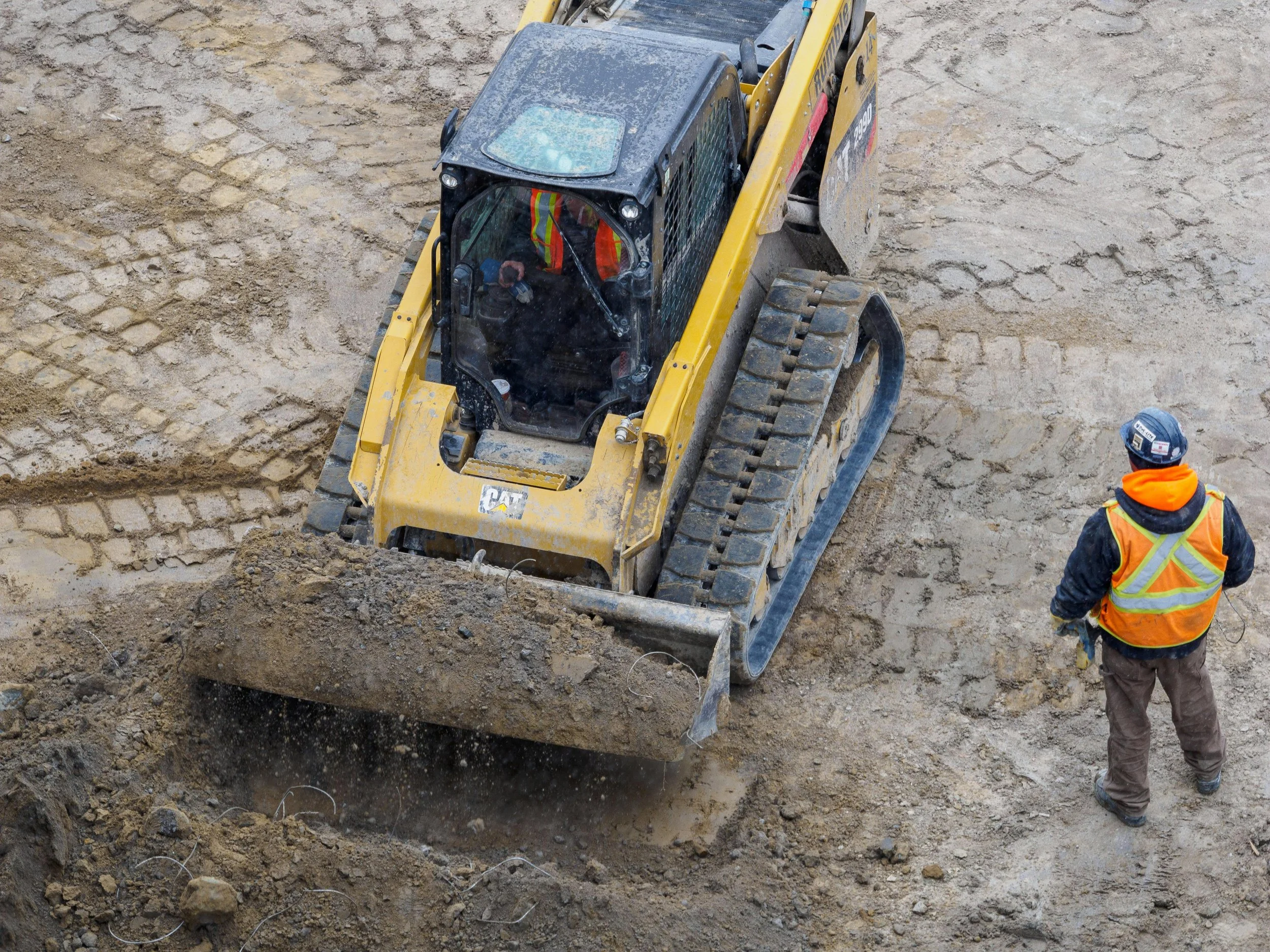 A construction worker wearing a safety vest and helmet operates a yellow bulldozer on a dirt construction site.