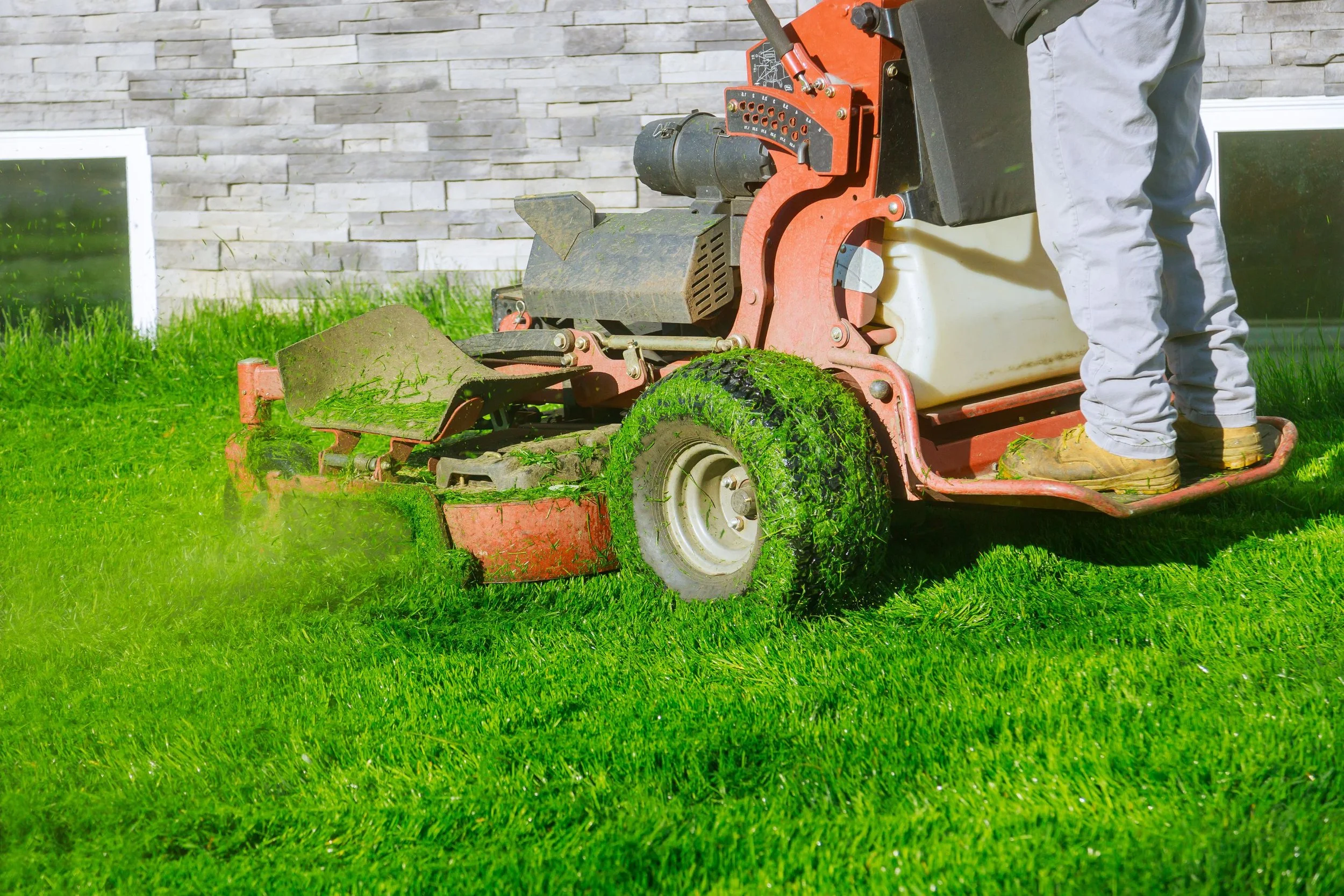Person operating a green lawn mower on a grassy yard with a stone wall and windows in the background.