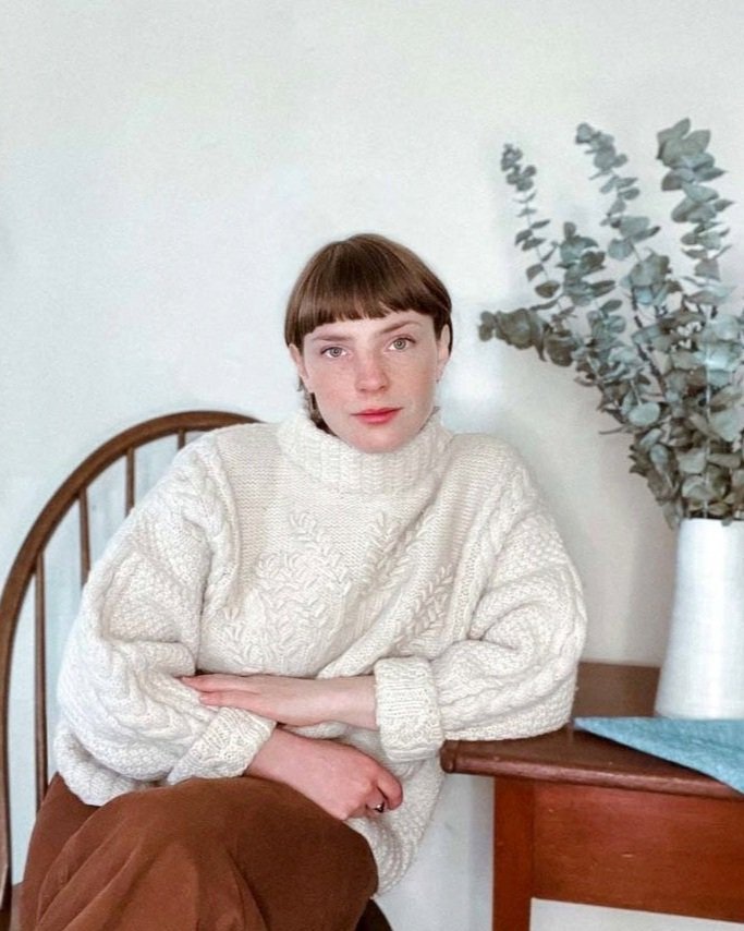 A woman with short brown hair and bangs, wearing a cream-colored knitted sweater, sitting at a wooden table with a blue cloth, next to a white vase with dried eucalyptus branches, in a room with light-colored walls.