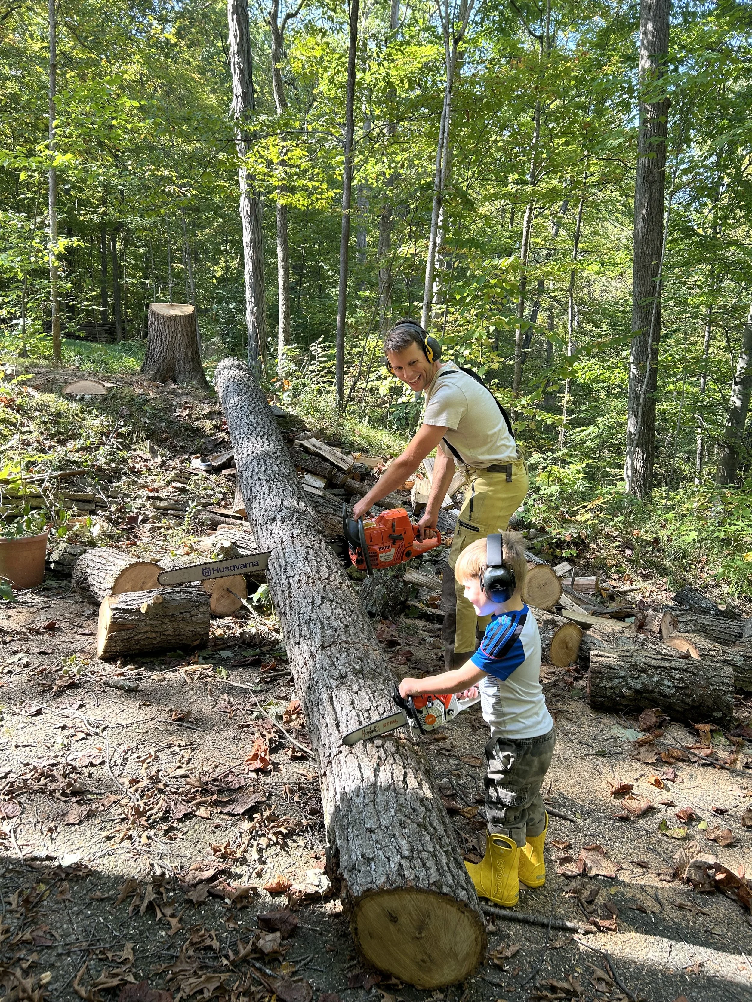A man and a young boy are cutting a fallen tree in a wooded area with chainsaws. The man is smiling and wearing hearing protection, while the boy is focused on using his chainsaw, both wearing protective gear including ear protection and boots.