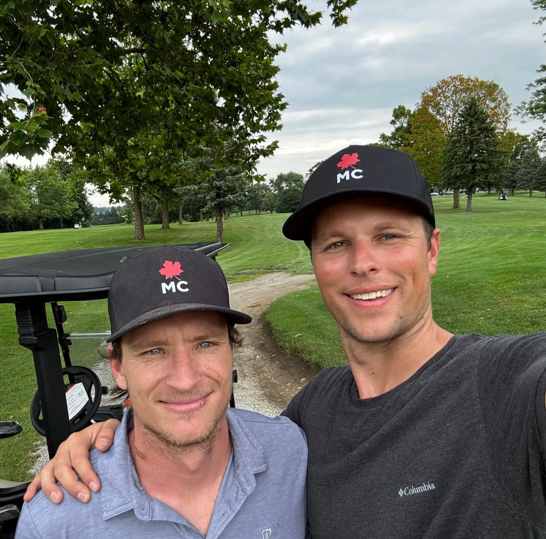 Two men taking a selfie on a golf course, both wearing black caps with a red maple leaf and 'MC' logo. One man has his arm around the other's shoulder.