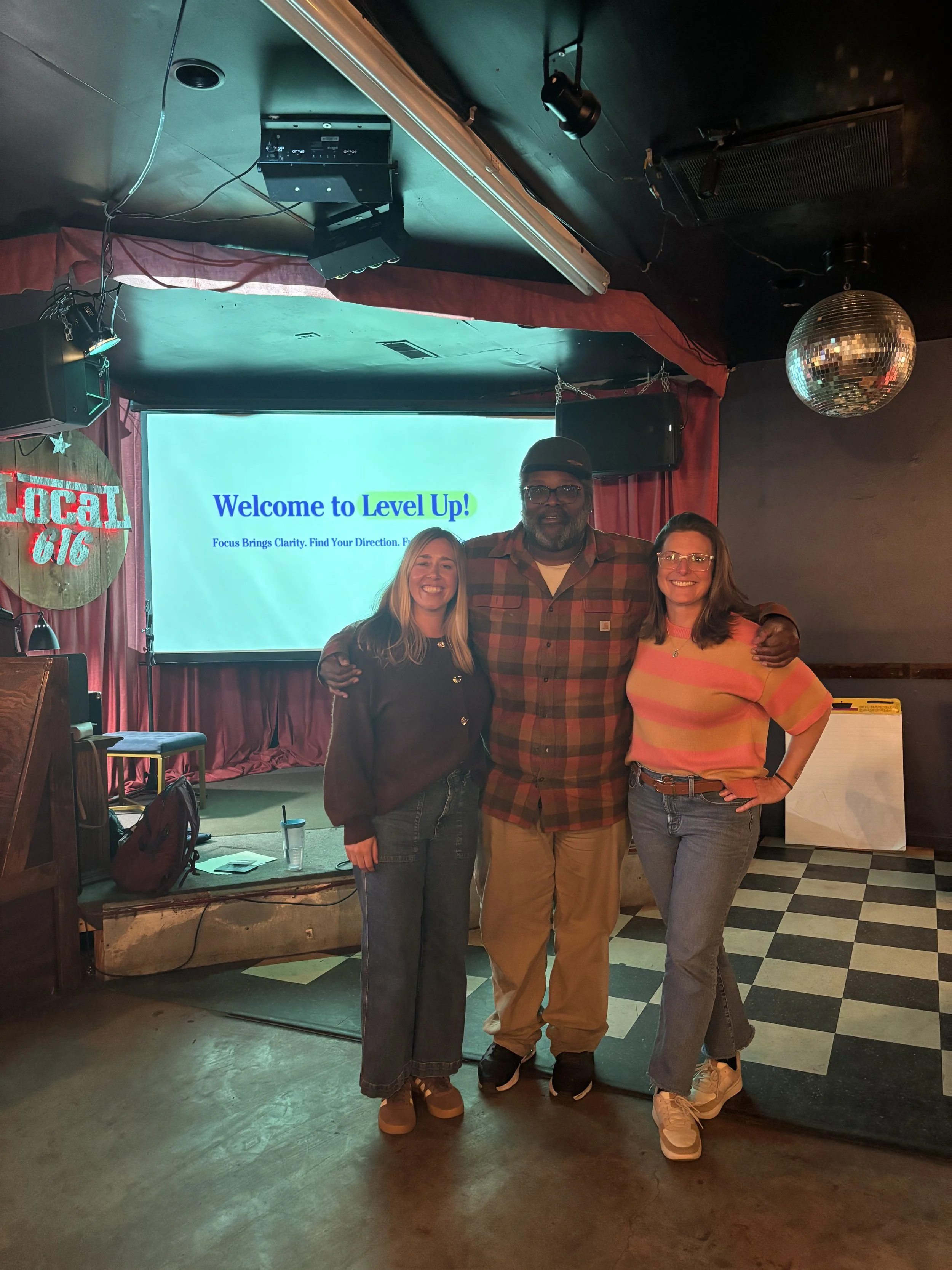 Three people standing together in front of a stage with a screen that reads 'Welcome to Level Up!' The stage has a backdrop with red curtains, a disco ball hanging from the ceiling, and a sign that says 'Local Gig' on the left.