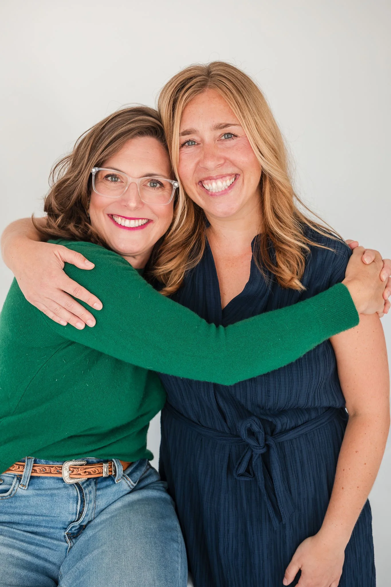 Two women hugging each other and smiling at the camera, one with glasses and the other with long blonde hair, dressed casually.