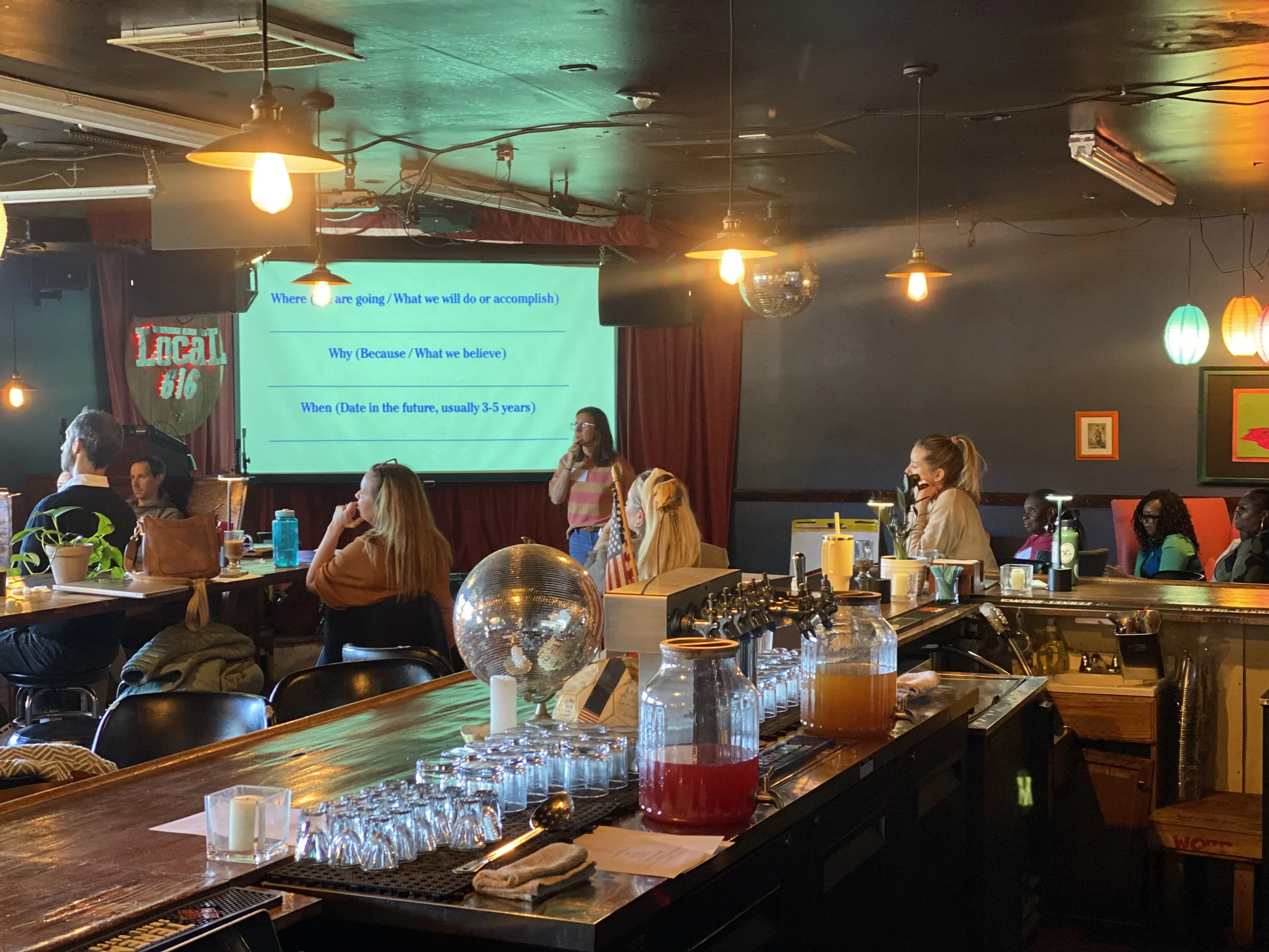 A woman is giving a presentation on a stage in a dimly lit bar or restaurant, with a large screen behind her displaying a slide with text. Several audience members are seated, listening, and the room has decorative lights and a bar with drinks and glasses in the foreground.