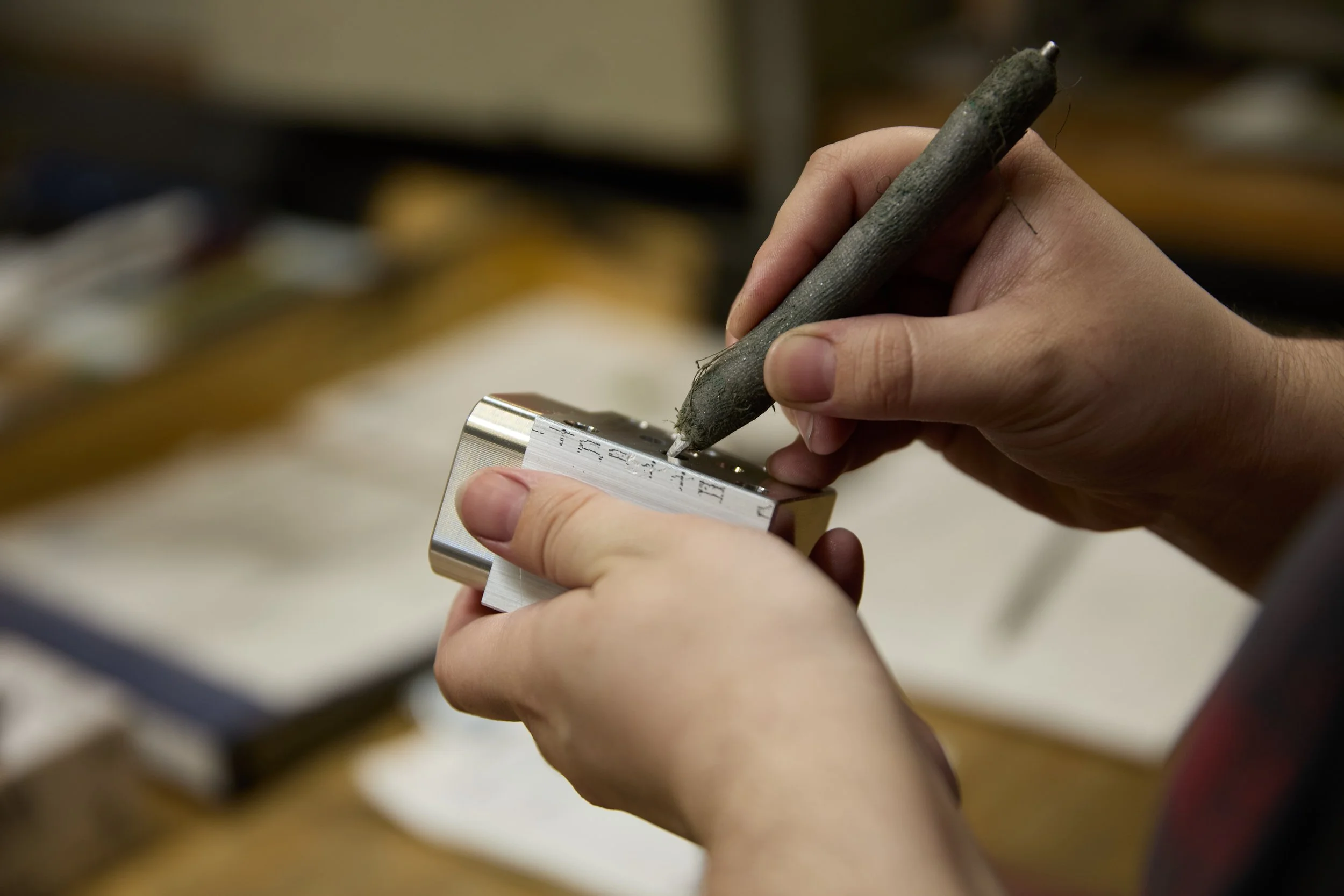 Person's hands carving or engraving a metal piece using a stylus and a metal block.