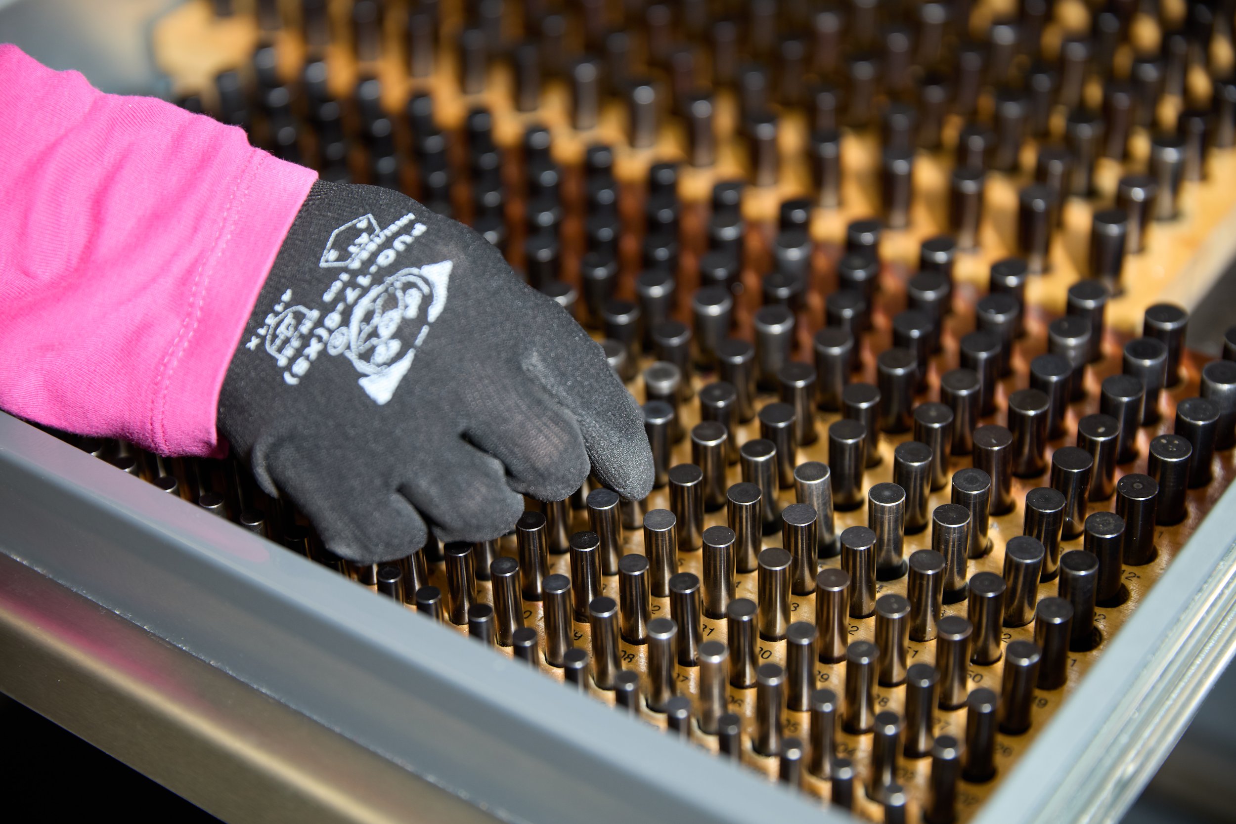 A worker wearing gloves handling a tray of small, black electronic components with numbered labels.