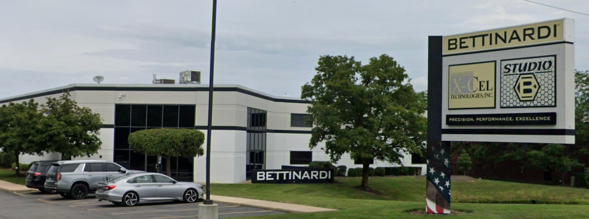 Exterior of a modern office building with a parking lot containing three cars. A large sign displays 'BETTINARDI', 'STUDIO', and logos for X-Cel Technologies Inc. The sign also has the tagline 'Precision. Performance. Excellence.' in smaller letters.