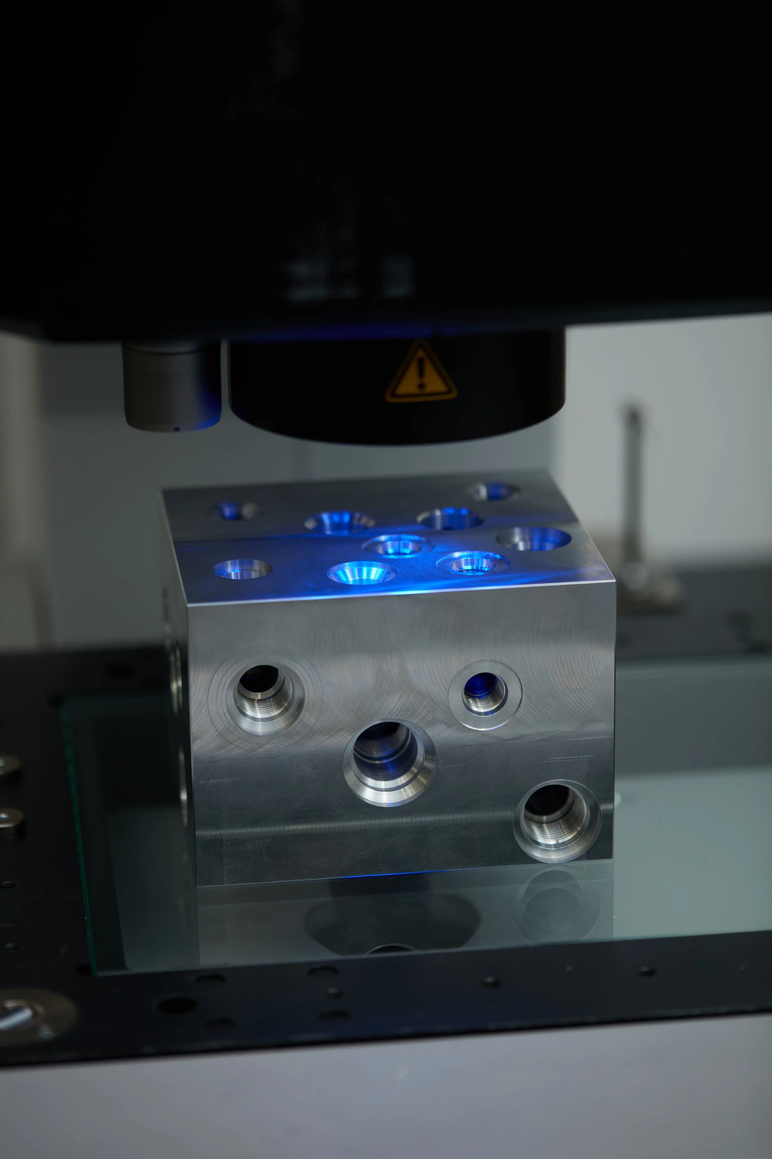 A close-up photograph of a metallic scientific or industrial device, possibly a microscope or precision instrument, with multiple circular holes and illuminated blue lights, in a laboratory setting.