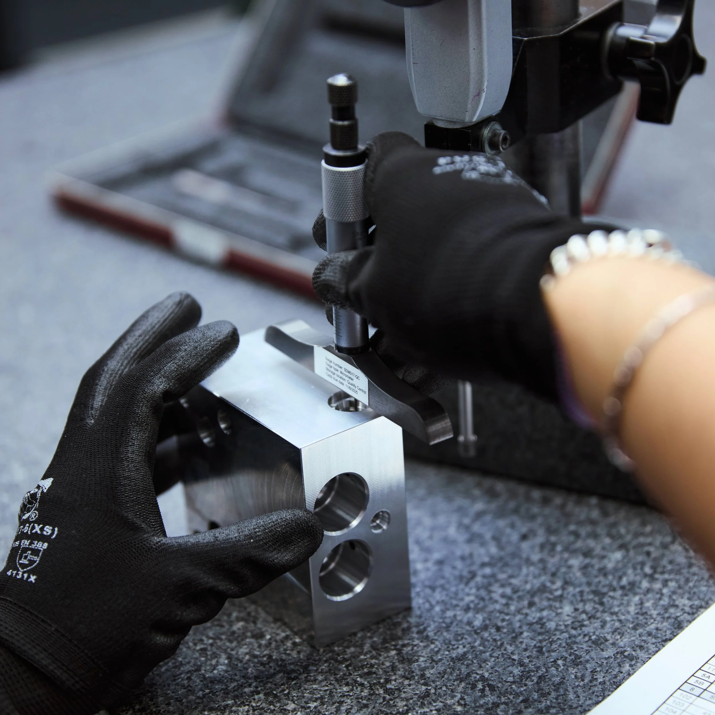 Worker wearing black gloves using a precision tool to machine a metal block with holes, on a workbench with a toolbox in the background.