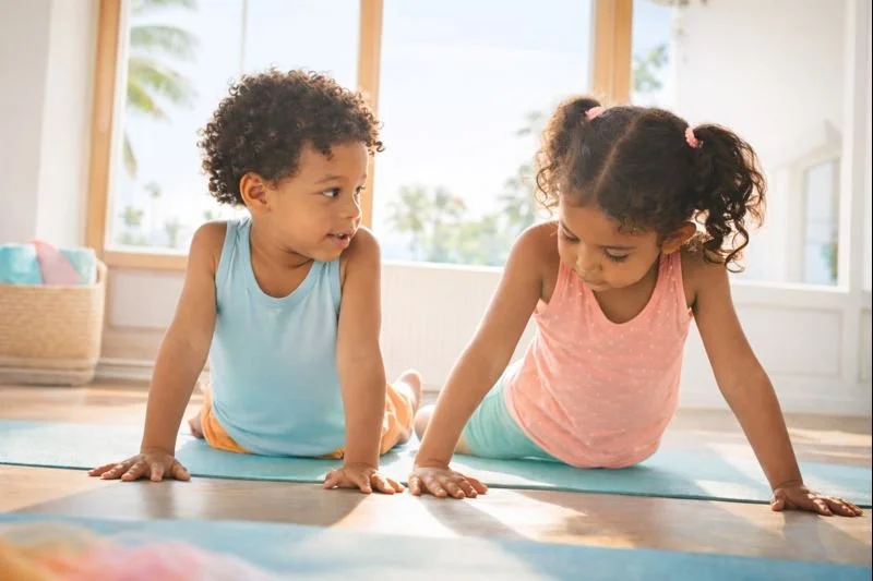 Two young children doing yoga or stretching on mats indoors near large windows