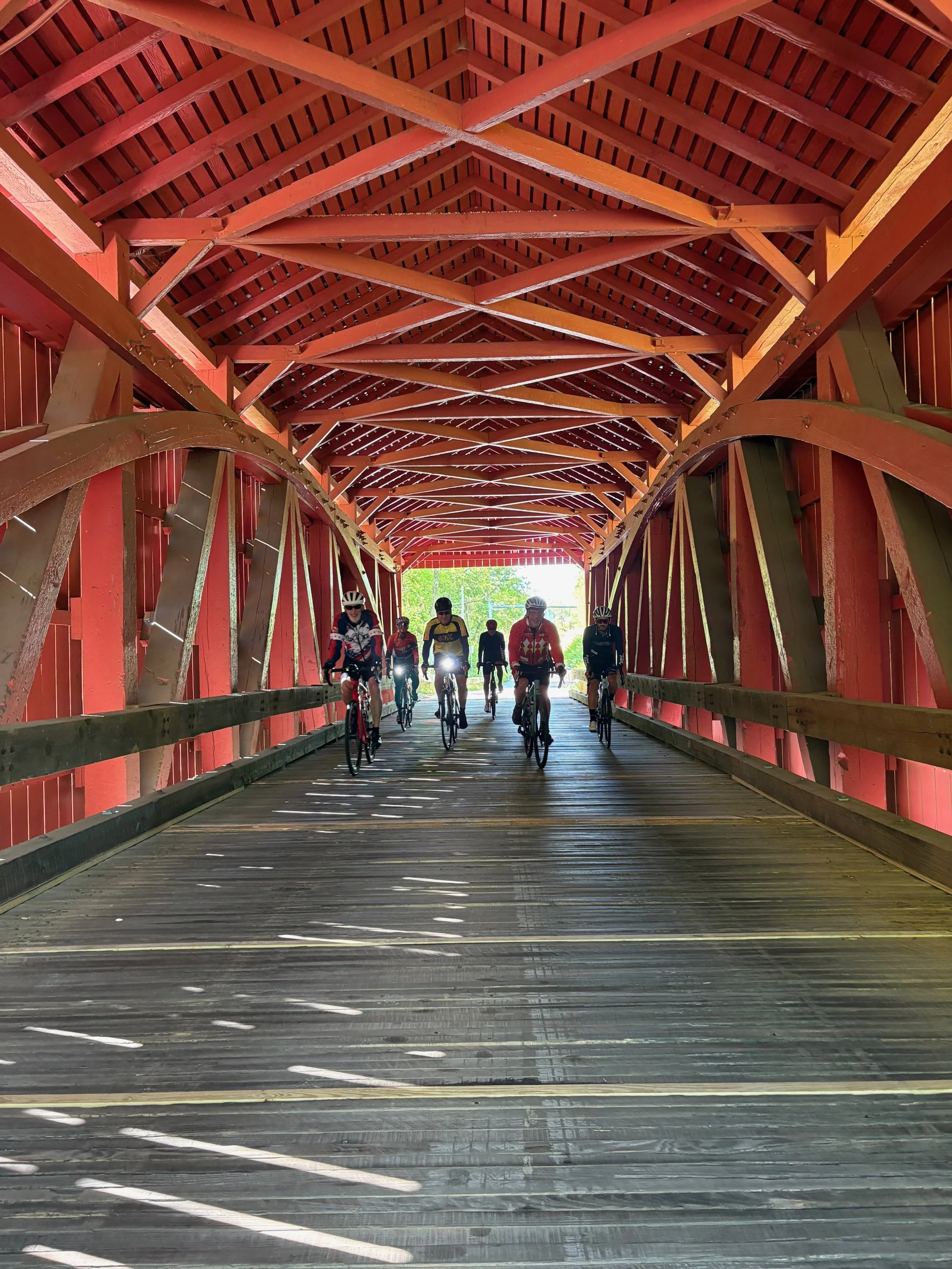 A group of cyclists riding through a red covered bridge with a wooden floor, visible sunlight, green trees outside.