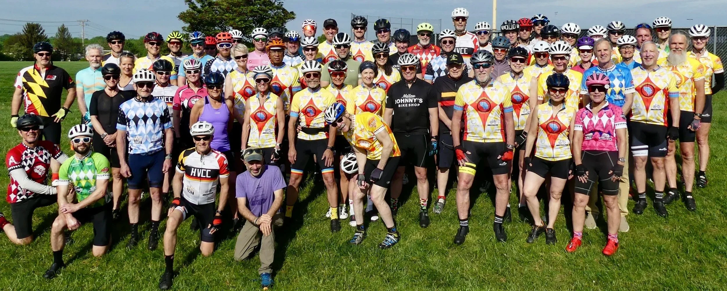A large group of cyclists wearing colorful jerseys and helmets pose together on a grassy field. Some are kneeling in front, while others stand behind. The background features a chain-link fence, trees, and a blue sky with clouds.
