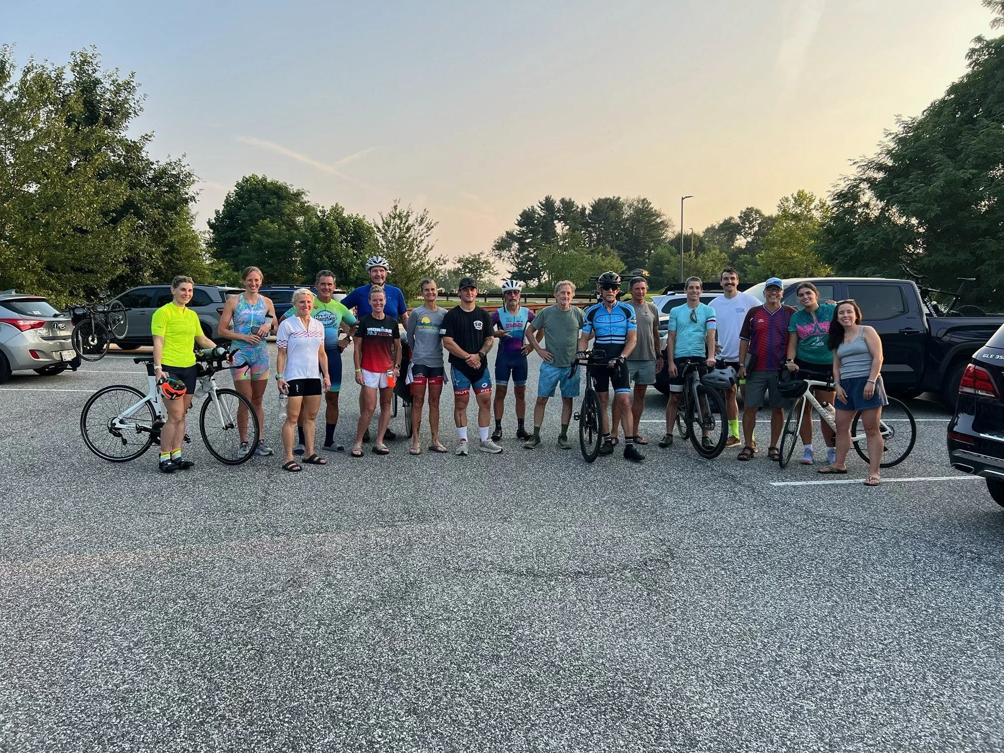Group of people standing in a parking lot, some with bicycles, during what appears to be a cycling event or ride, with trees and parked cars in the background at sunset.