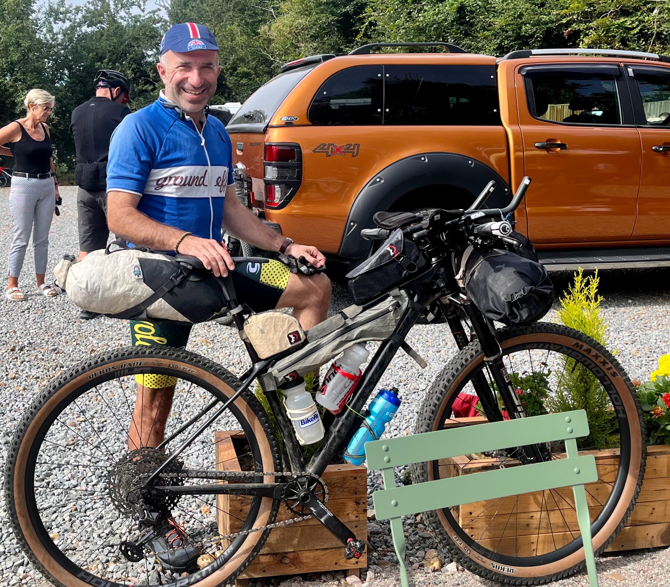 A man in cycling gear sitting on a mountain bike, smiling at the camera, with a brown truck and other people in the background.