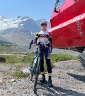 Person standing with a bicycle outdoors near a large red structure, with mountains in the background